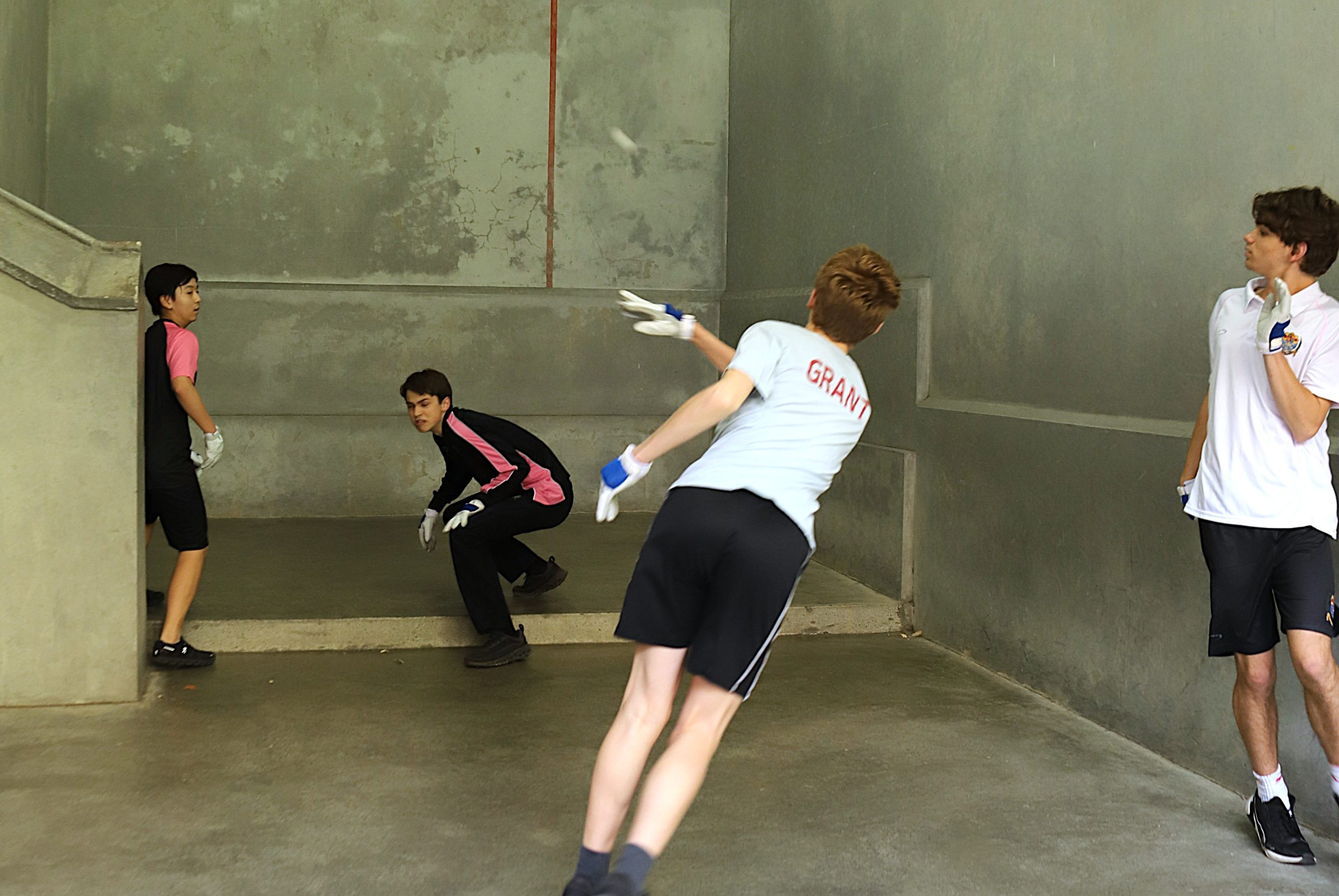 Four boys playing Fives on the Fives court, one boy has hit the ball mid air and another boy has ducked
