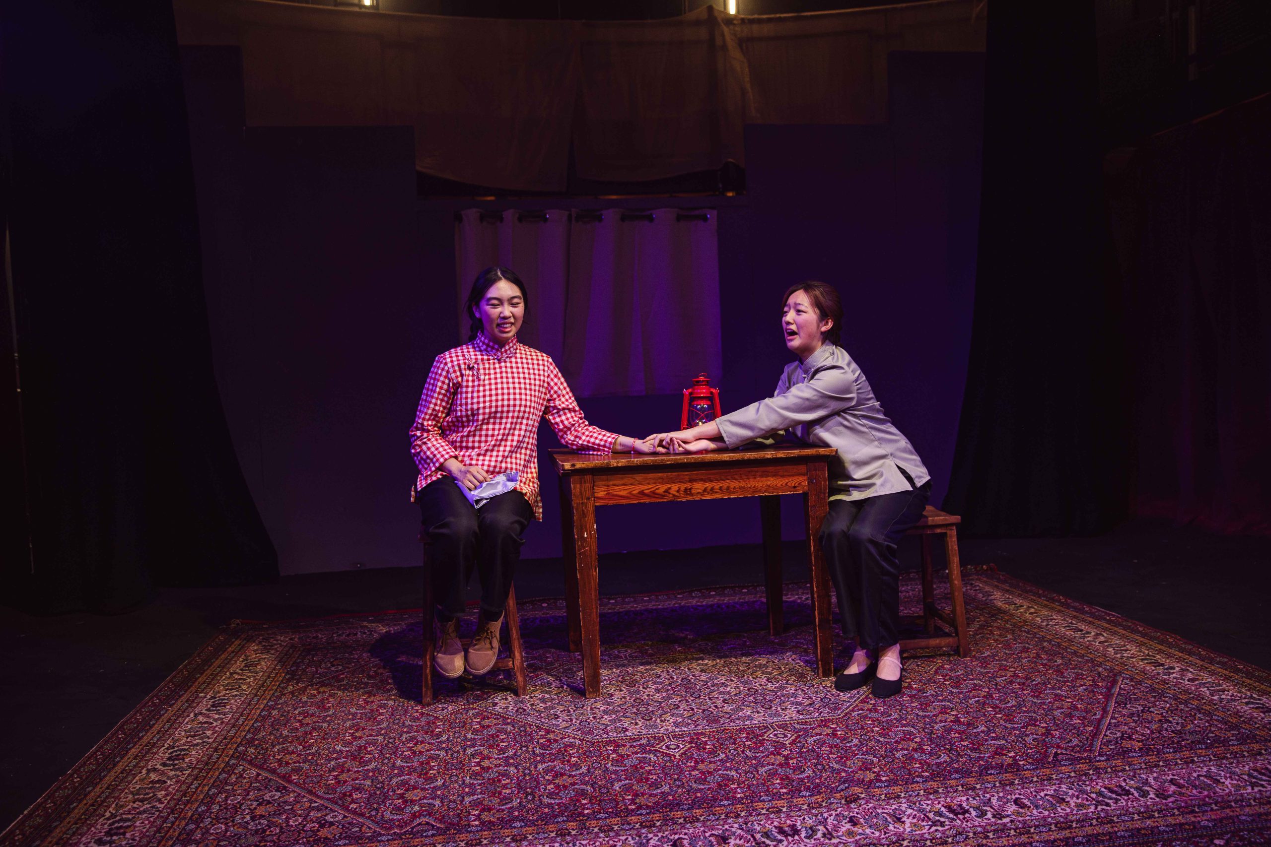 Two girls on stage, seated at a table, one holding the other's hand