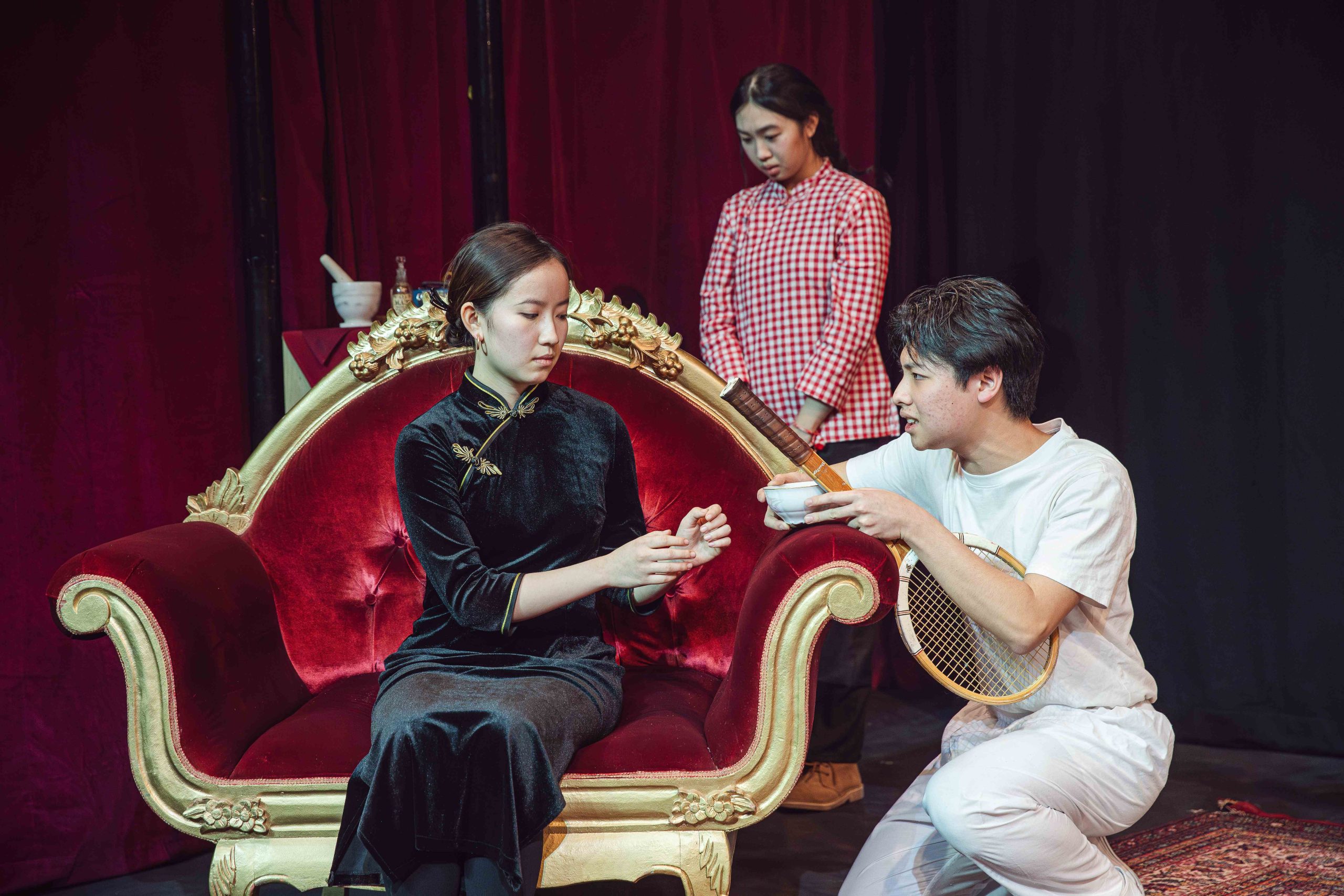 Two girls and a boy on stage, one girl is sat down and the boy is offering her tea