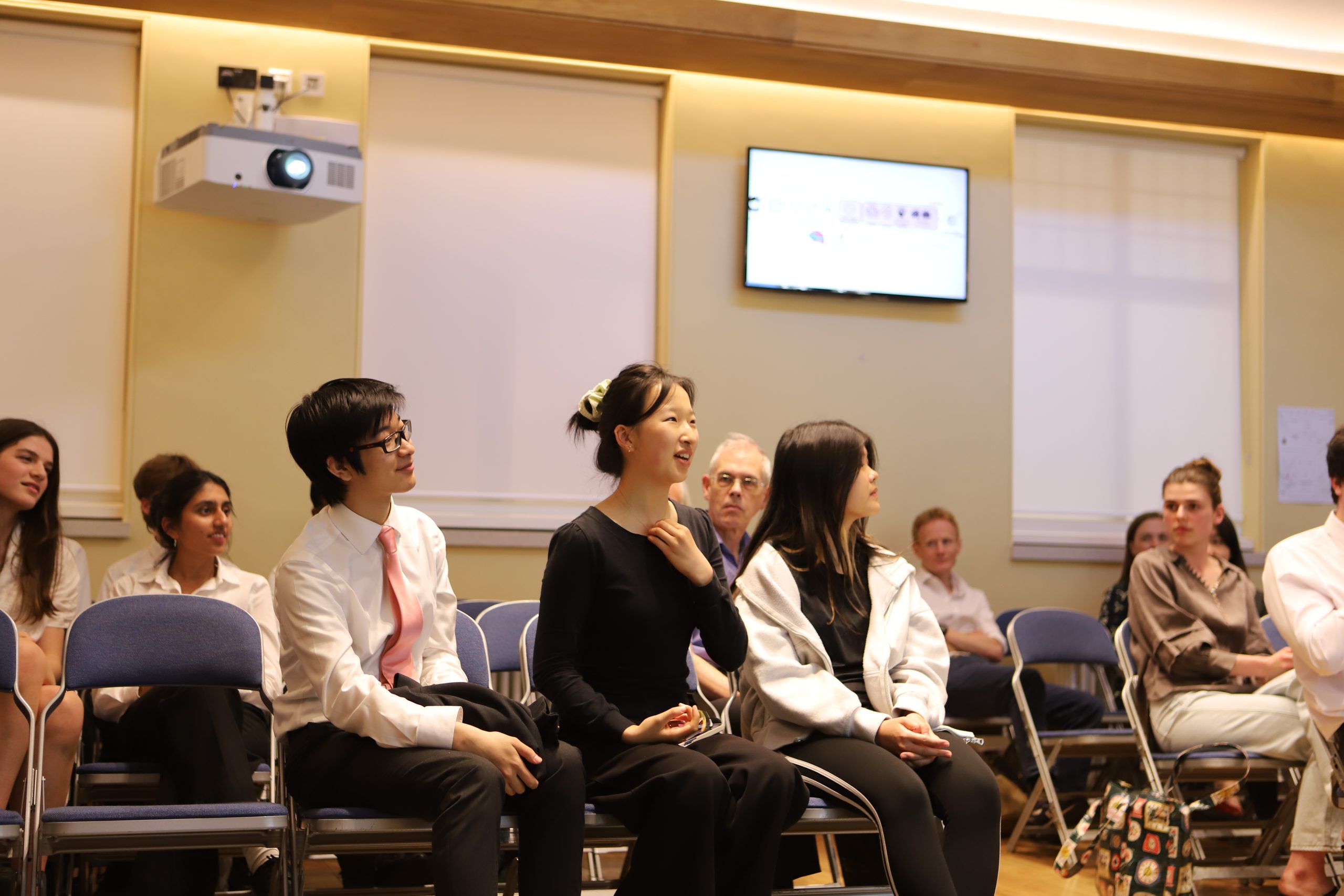 Three pupils, one male and two female. On the front row. The girl in the middle is asking a question.