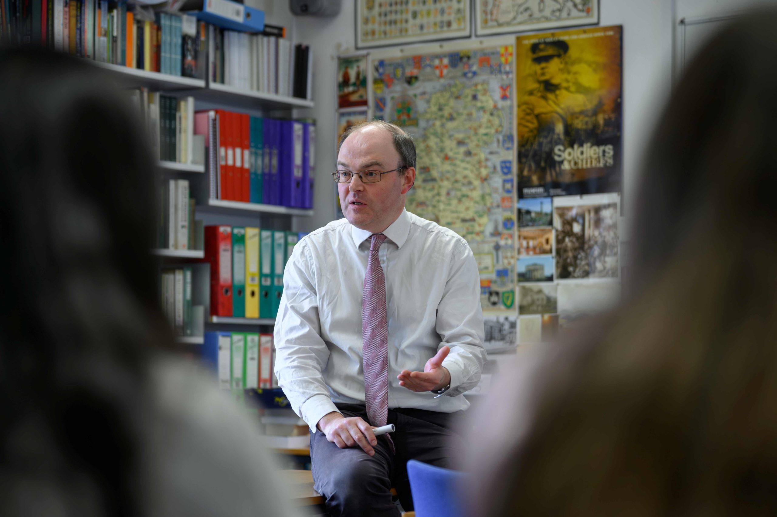 A male teacher sat on his desk teaching, with folders and maps in the background and the silhouettes of a pupil either side