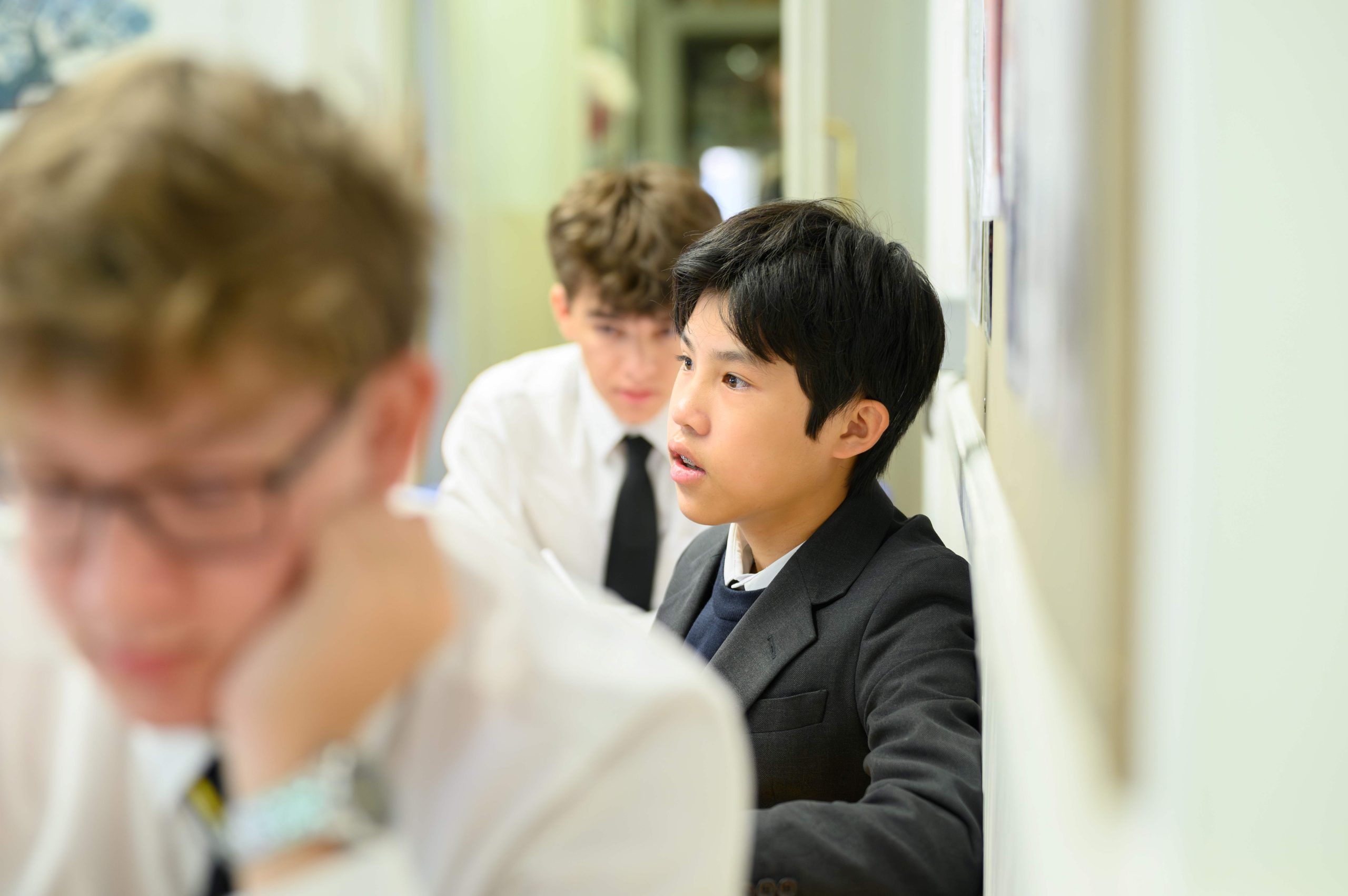 A boy looking towards the front of the class, sat with his back to the wall, with another boy behind him