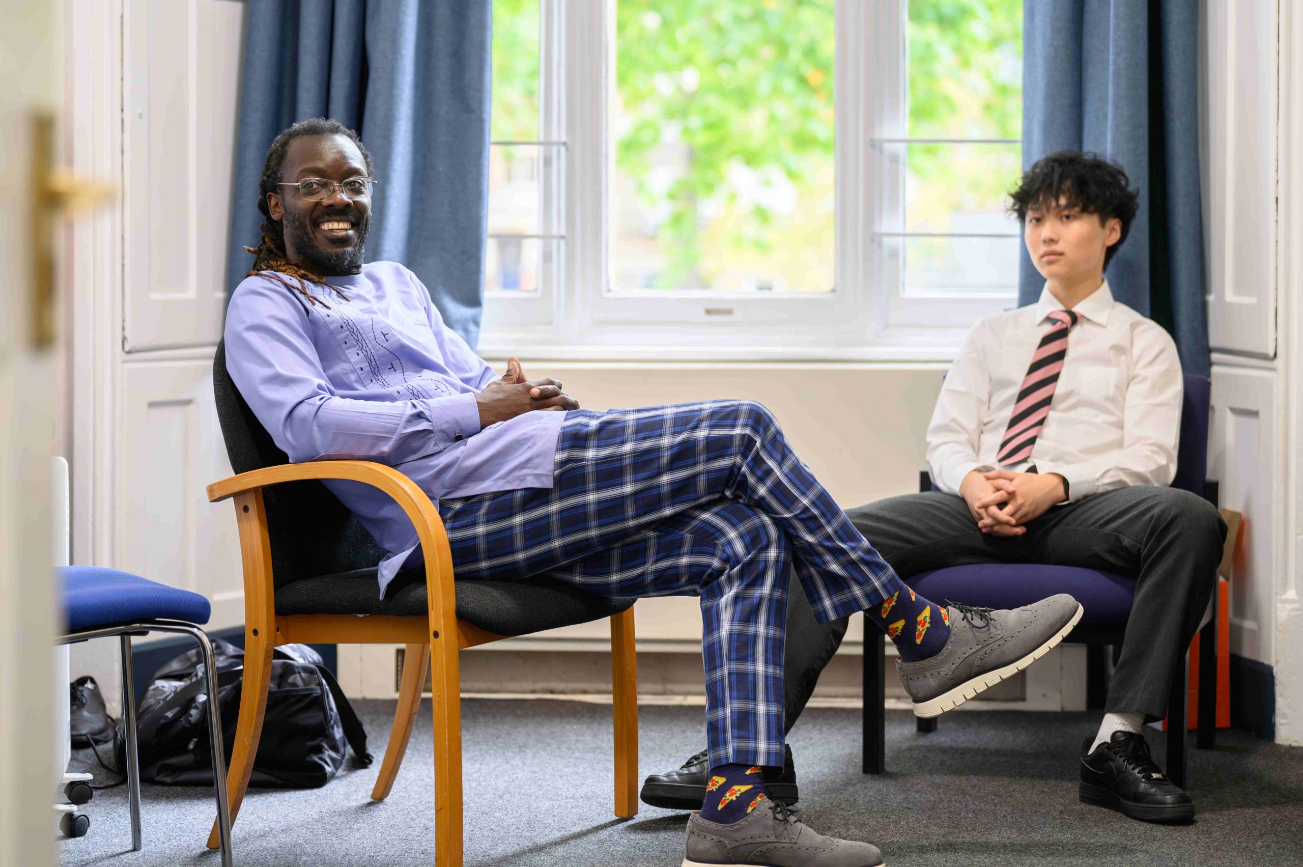 A housemaster sat talking with a pupil in his dorm, in front of the window