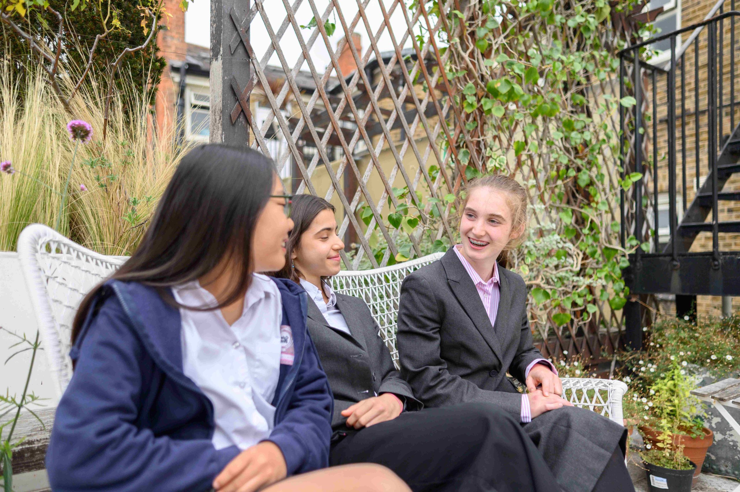 Three girls sat on a bench together on a roof terrace, with a trellis and plants behind them