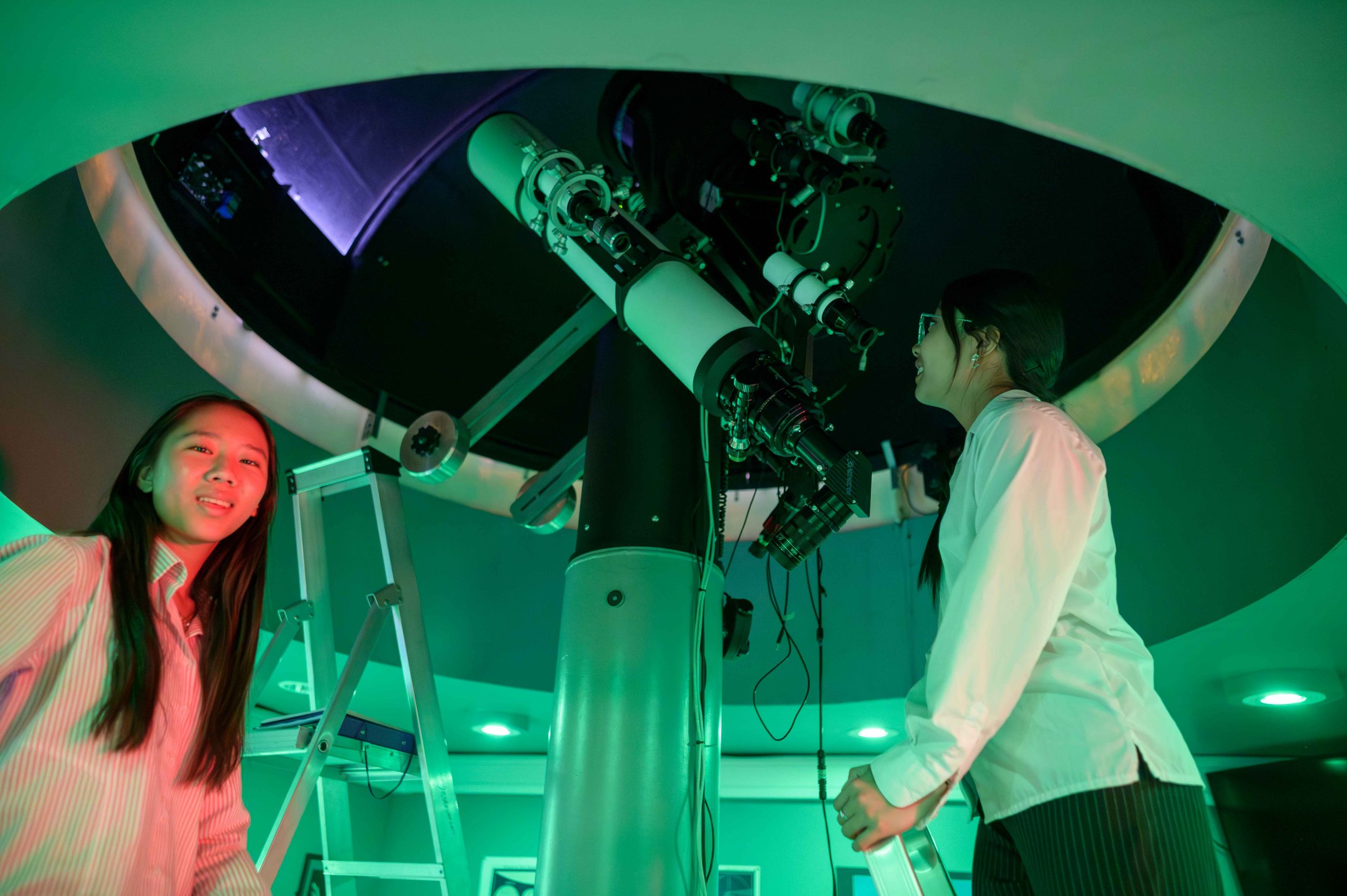 A girl looking into the telescope in the School's observatory, with another girl smiling at the camera
