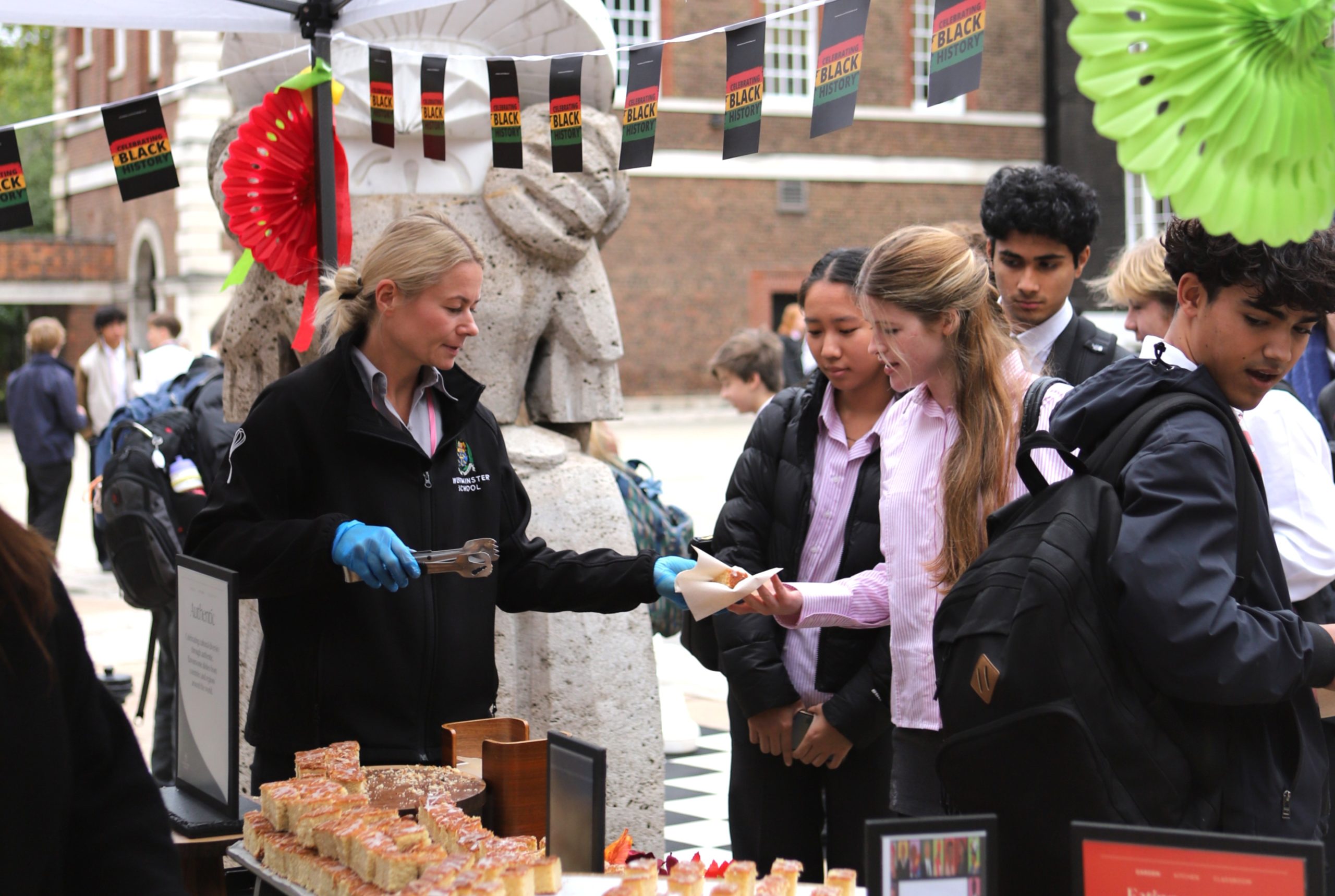 Member of the catering team handing out Jamaican spiced cake to two Sixth Form girls at the front of the queue, at the BHM pop-up stall in Little Dean's Yard.