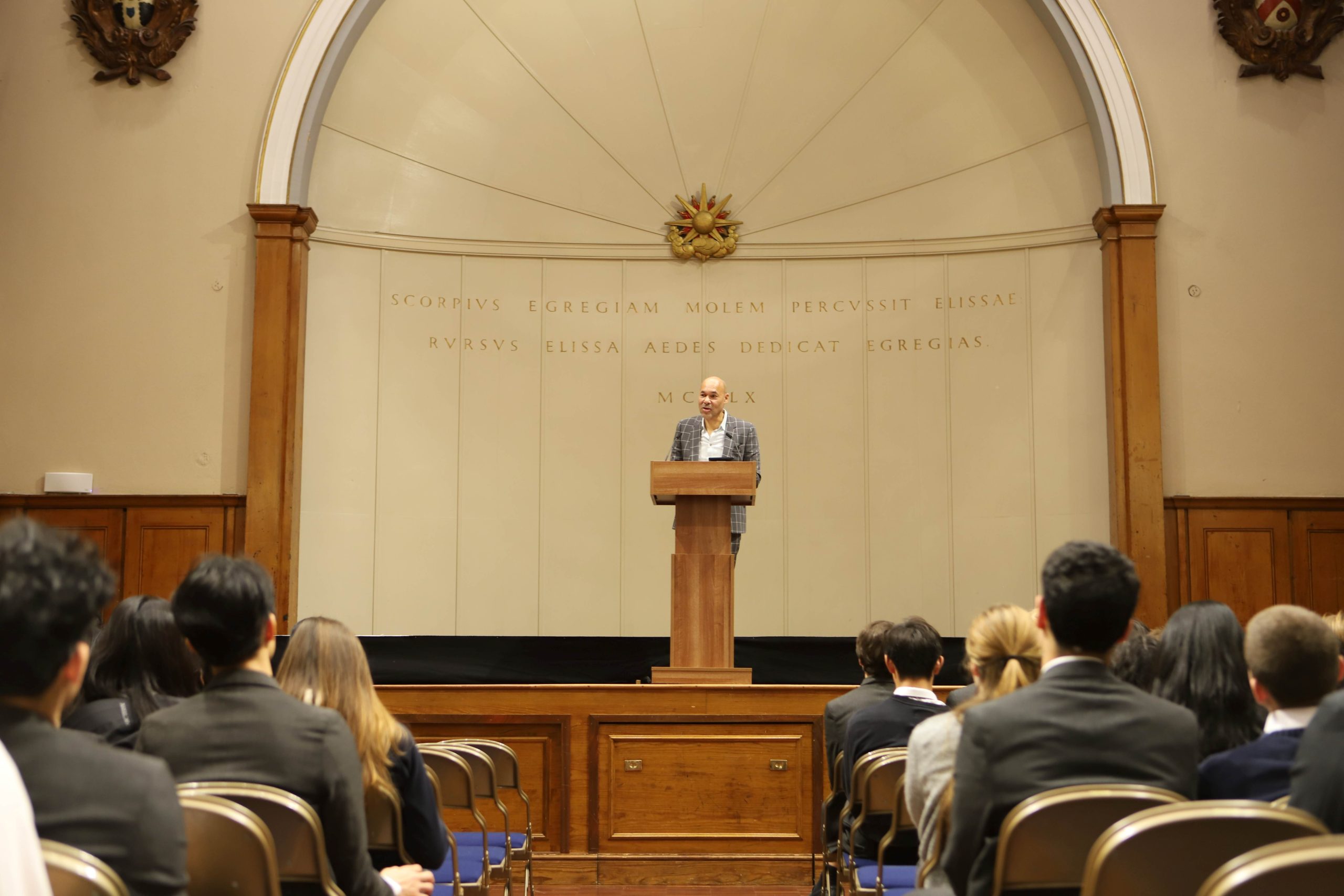 Basi speaking on stage in front of a lectern at a Locke lecture, with the audience in front