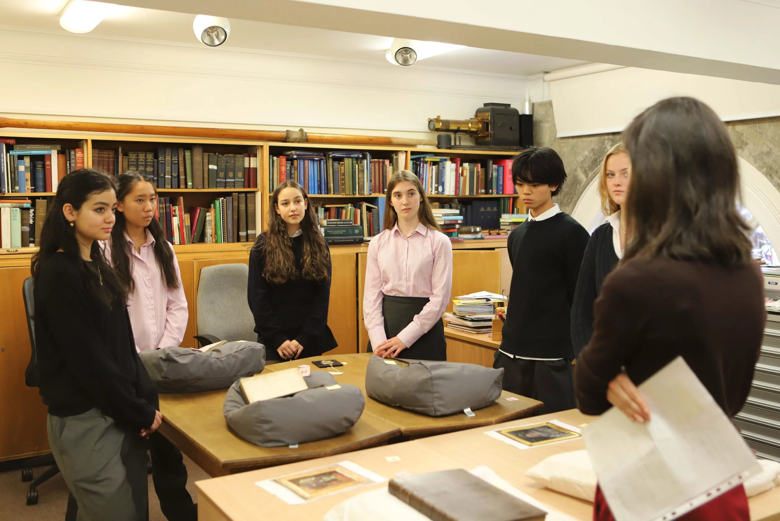 Four girls and a boy in a row around a table looking at artefacts, with the archivist at the front speaking to them and shelves of books behind them