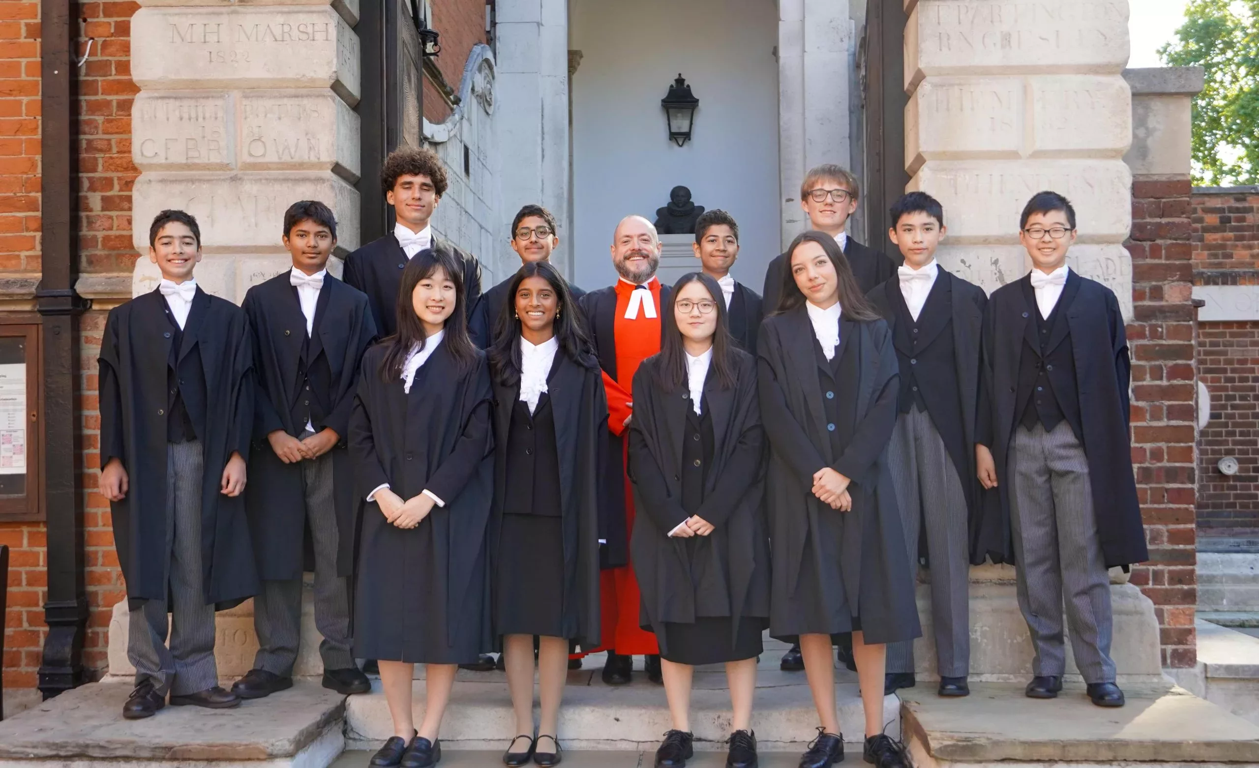 The King's Scholars 2025 standing in their black robes with their housemaster in red in the middle on the steps up to School from Little Dean's Yard with either side of the arch in view and a bust and lantern in the middle behind them