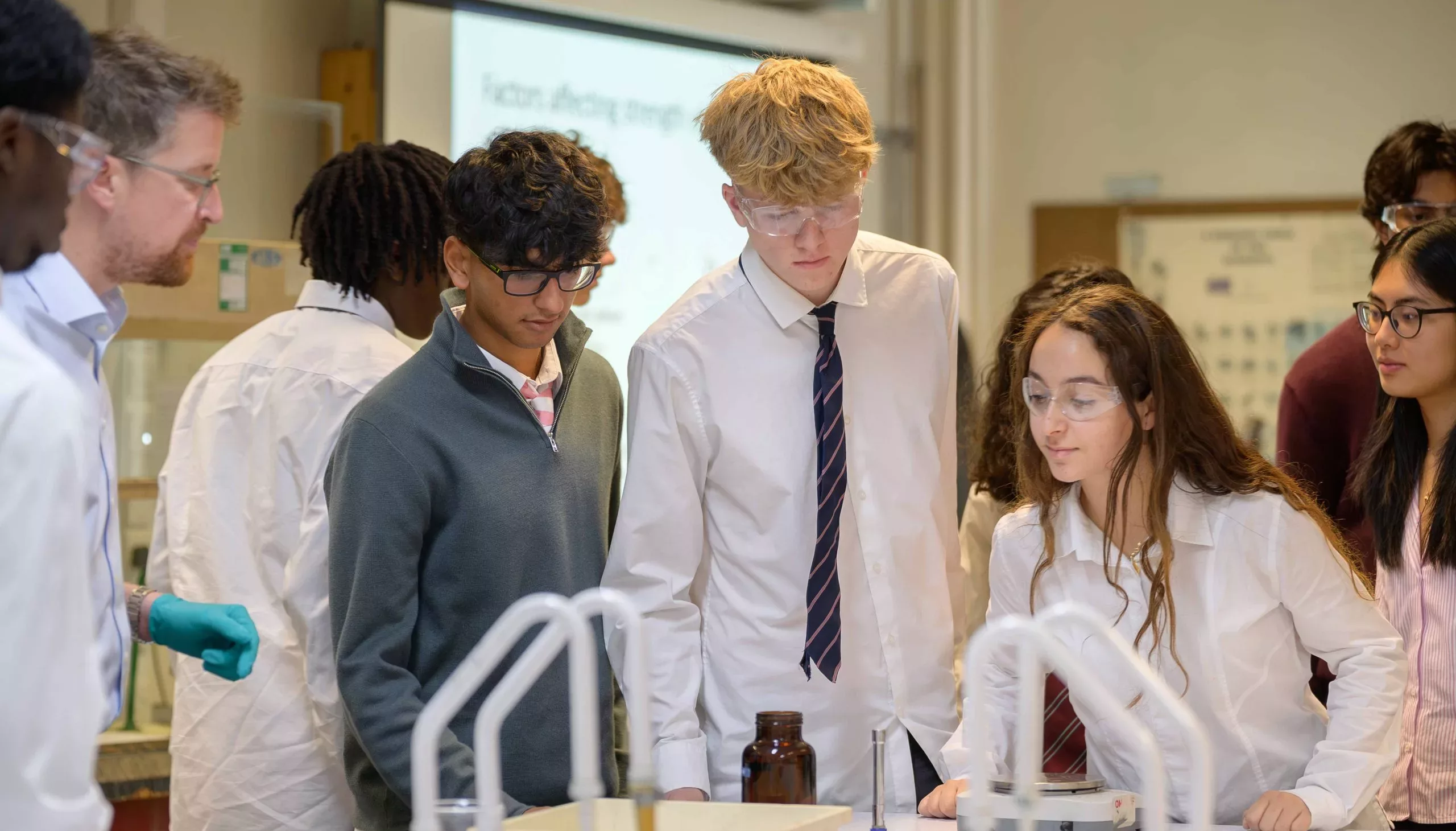 A group of boys and girls wearing googles in a Chemistry lab, looking at a bottle filled with something for their practical