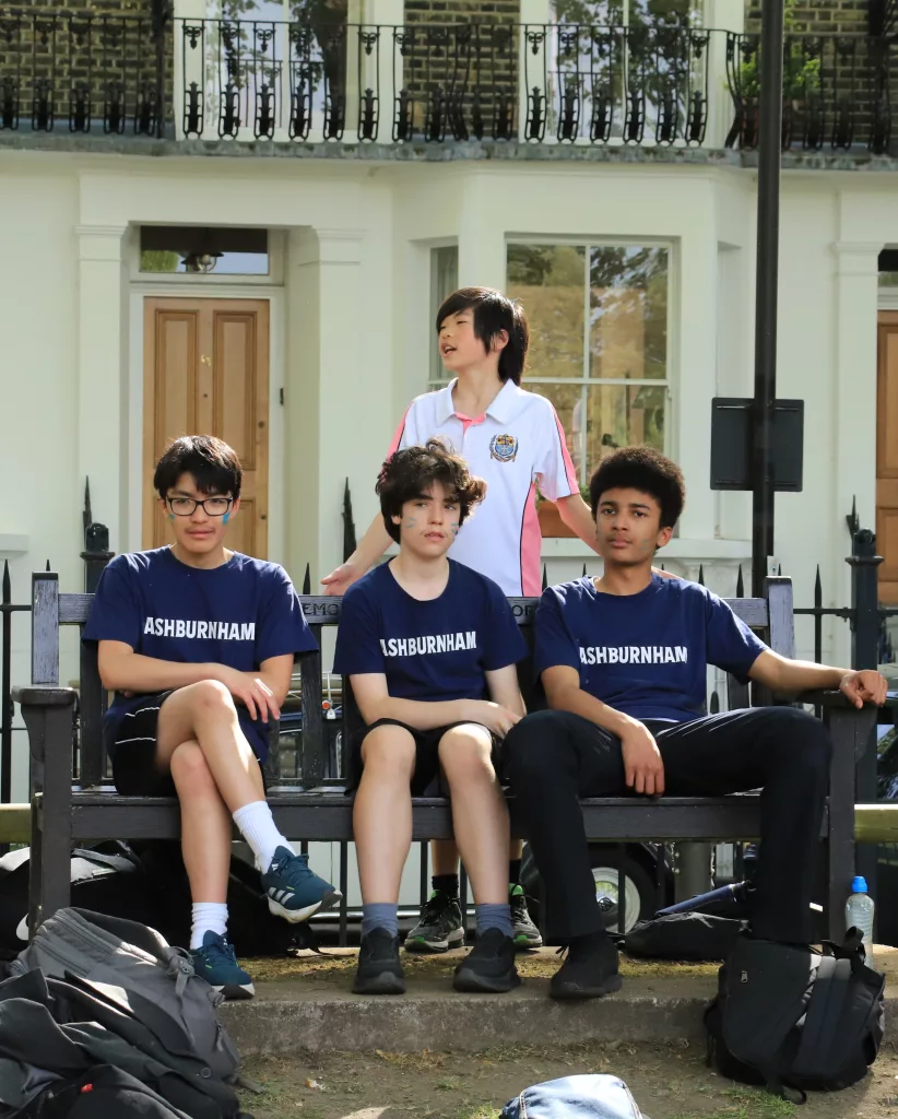 Three Ashburnham boys in navy house t-shirts sitting on a bench with a boy standing behind them in a Westminster sports top