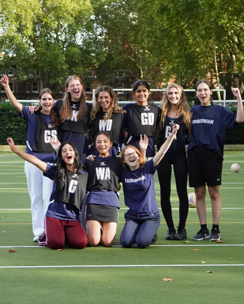 The Ashburnham girls netball team, six girls standing and three kneeling in front, in house t-shirts gathered for a football to celebrate their win at Vincent Square with their arms in the air