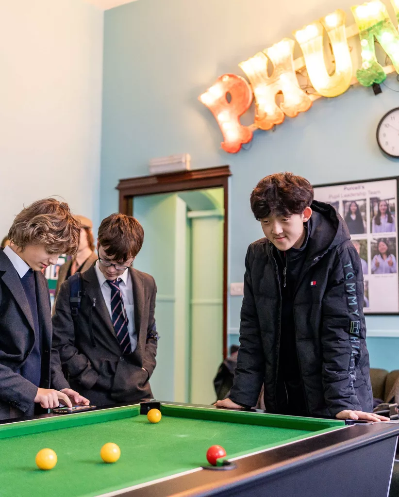 Three boys standing at Purcell's Common Room pool table with a lit-up sign behind them