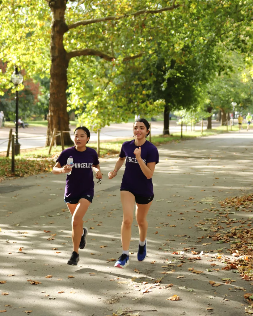 Two girls running towards the camera in their house t-shirts in a race in Hyde Park down a tree-strewn avenue