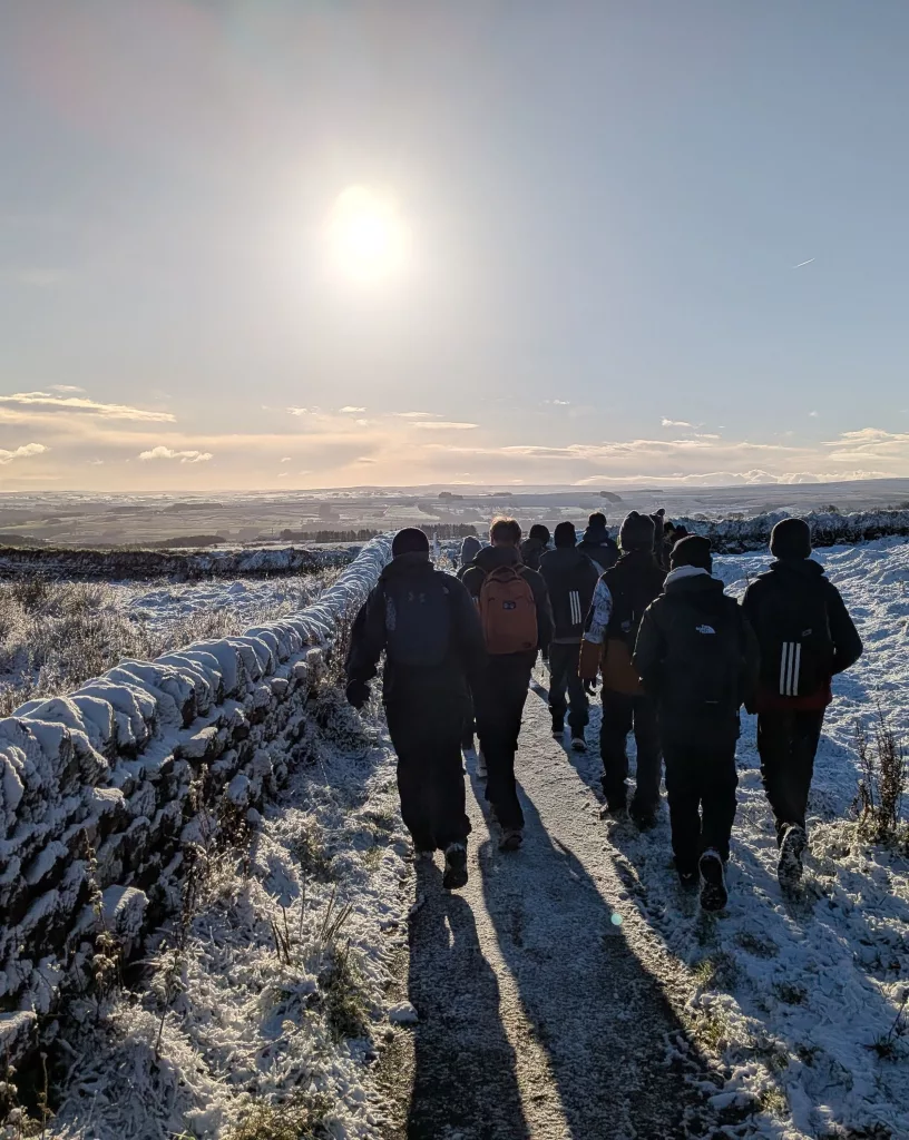 A group of boys walking down a snowy lane with dry stone walls either side, taking from behind with the sun high in the light blue sky