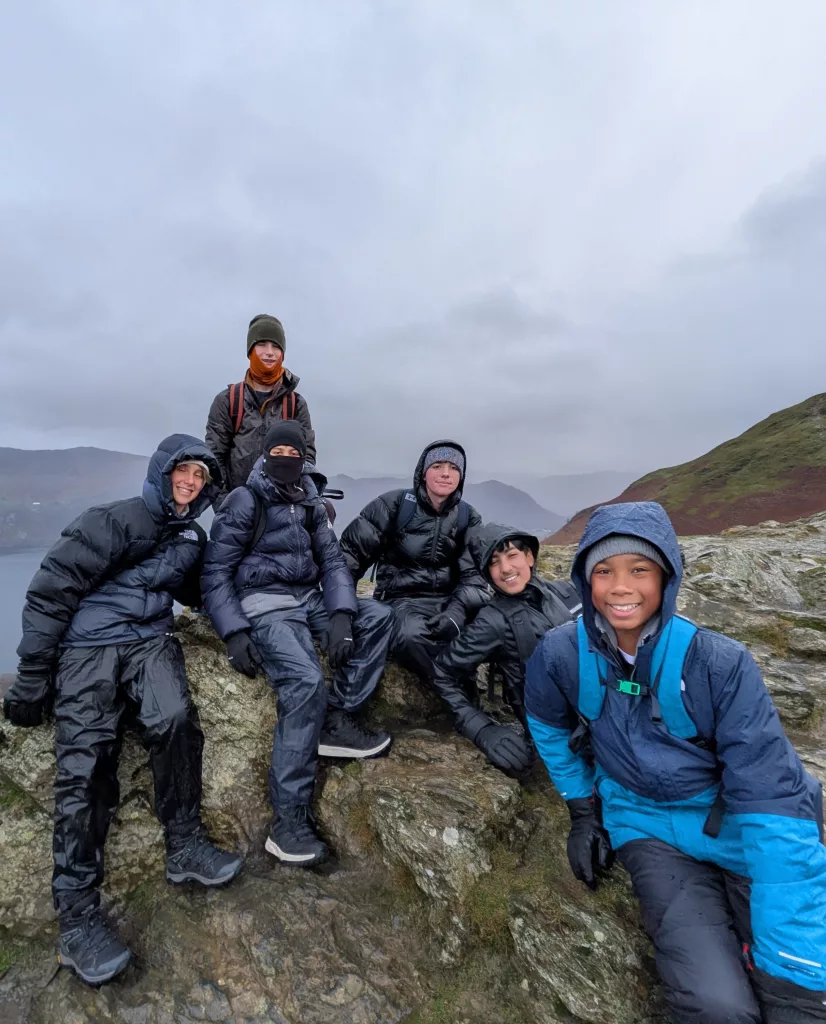 A group of boys in hiking gear posing for a photo slumped atop a mountain with a lake and more mountains in the background and a grey, cloudy sky