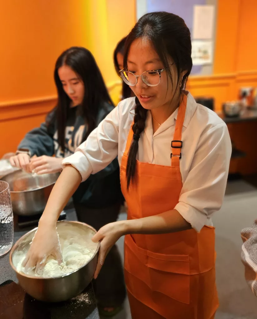 Two girls making dough, the one in front is kneading the mix in an apron on and the girl behind is handling dough