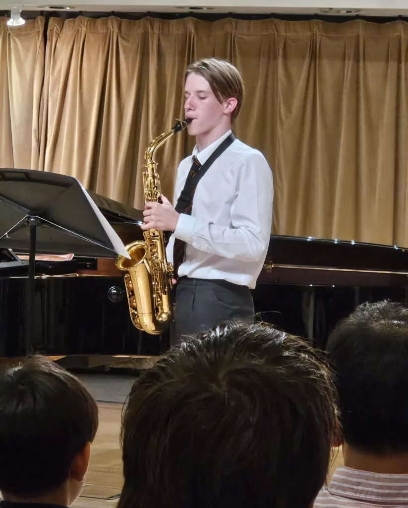 A boy playing the saxophone in school uniform with the back of three audience members in the foreground and a gold curtain behind him