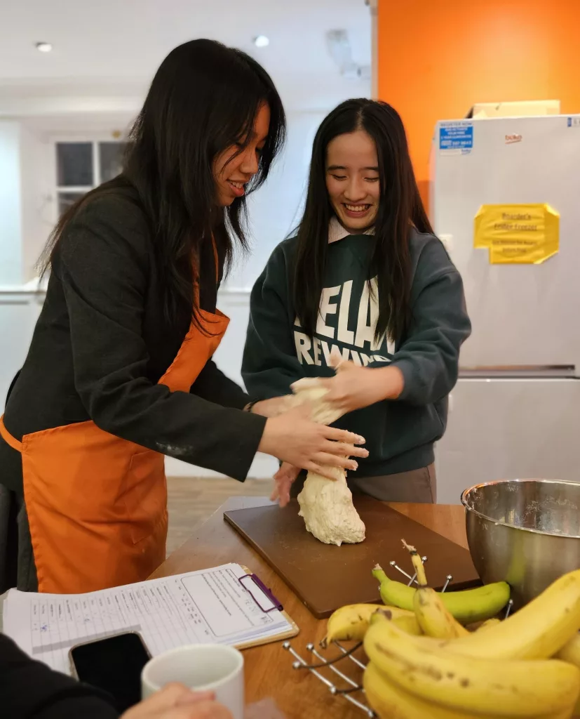 Two girls kneading dough on a wooden board, the one on the left is wearing an orange apron