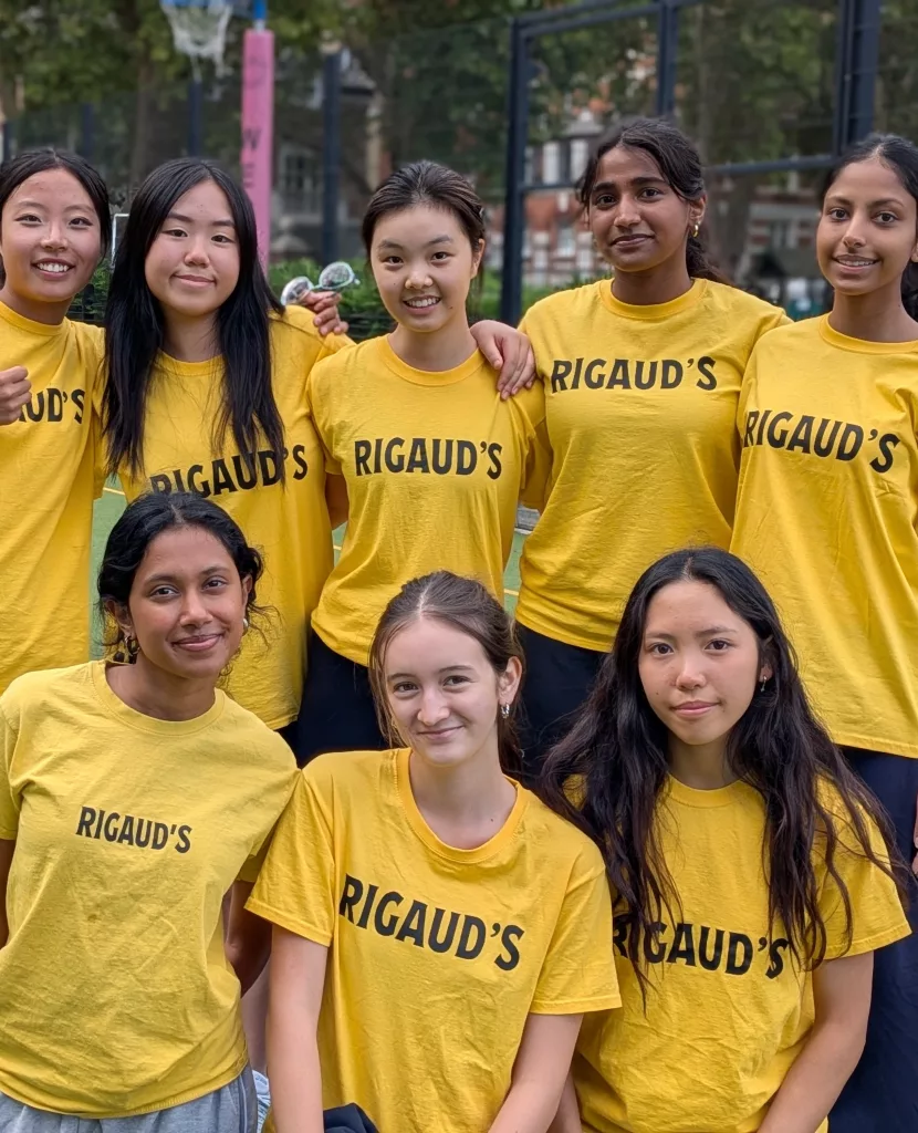 Five girls standing and three kneeling in front of them, huddled together for a group photo in yellow house sports t-shirts with a netball goal, trees, redbrick houses and fencing in the background
