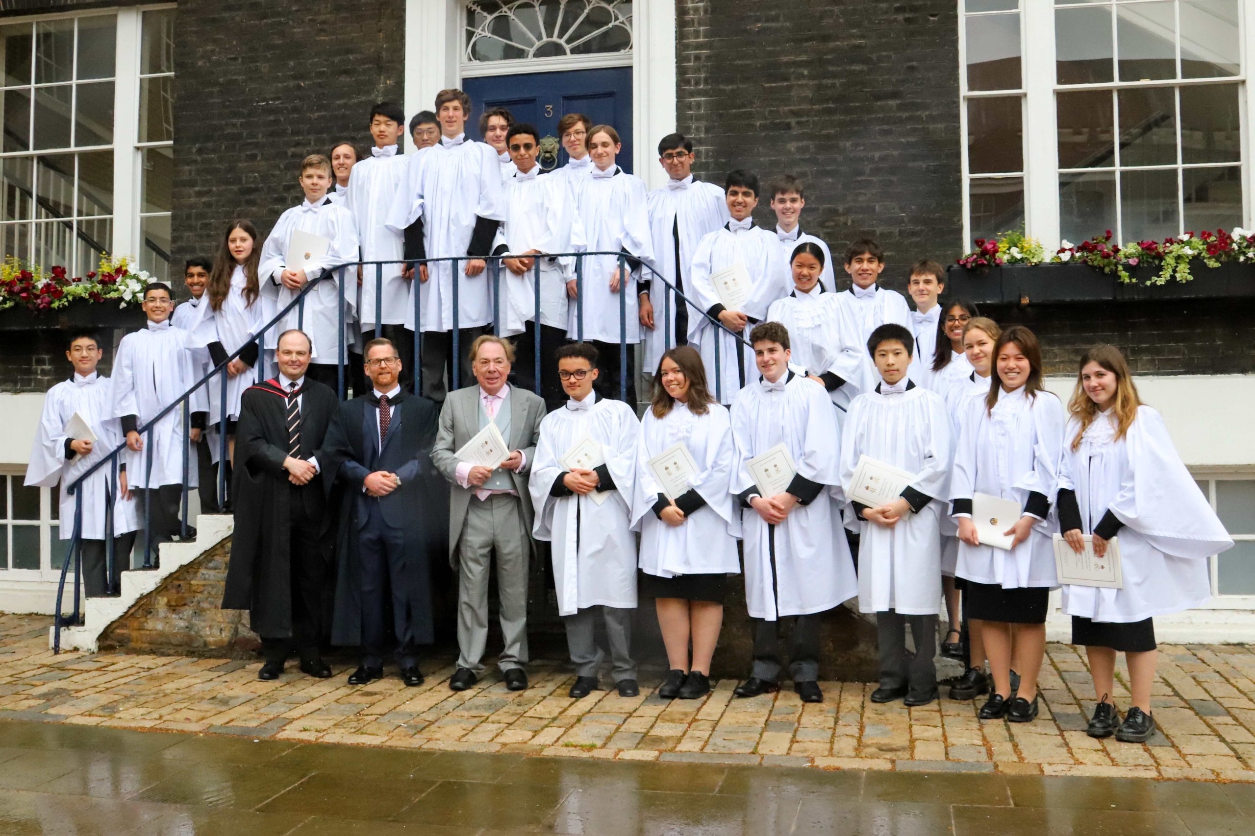 The King's Scholars lined up in their white robe on some steps up to a big blue front door on a Georgian town house - the boys and girls are standing on the steps either side of the door and on the stone in front in a line, with two gentlemen in robes and suits far left and Lloyd Webber between them and a male pupil in grey tails and pink shirt holding an A4 progamme and smiling