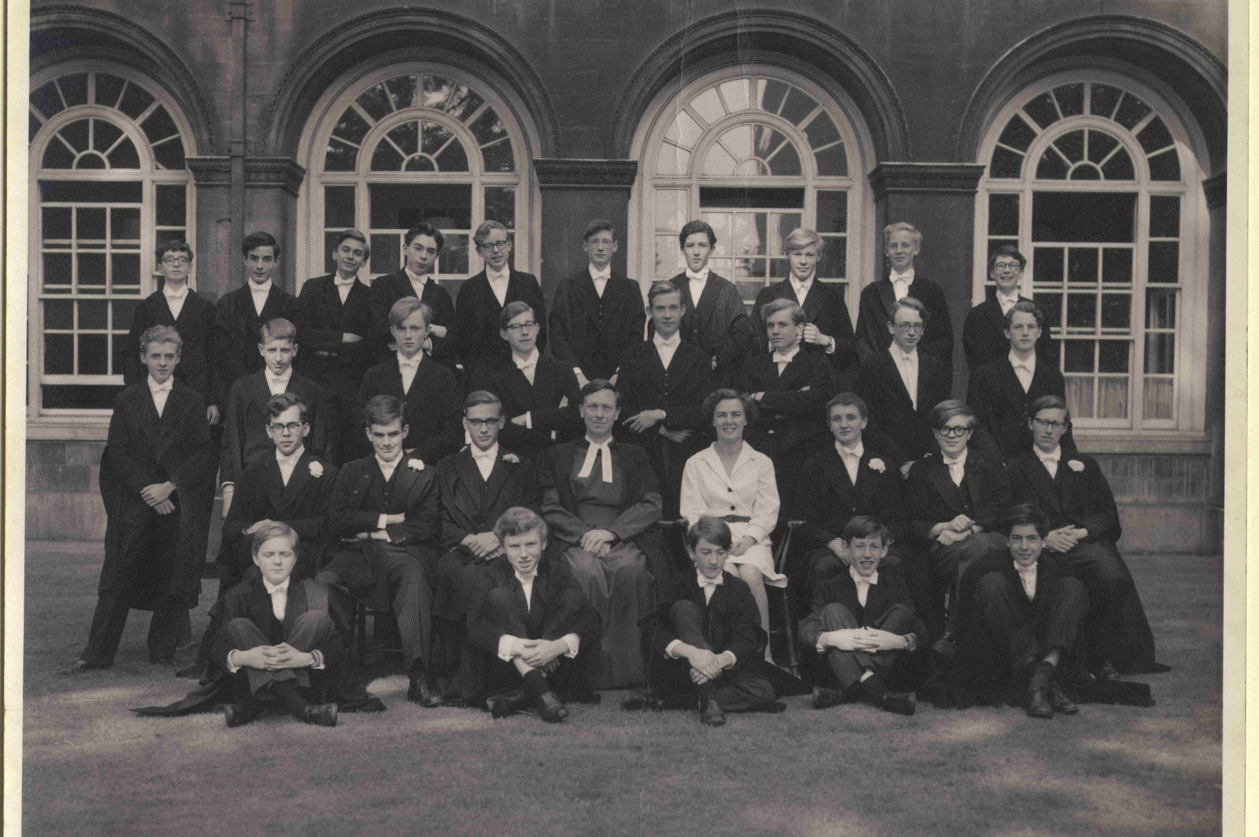A black-and-white photo of the scholars from Lloyd Webber's school days - they are in four rows wearing black robes and there is a male housemaster and a female teacher or matron in white in the second row - Lloyd Webber is in the far row, four in from the left and cocking his head jauntily, there are four arched crittall windows behind them with white joinery set in a stone building