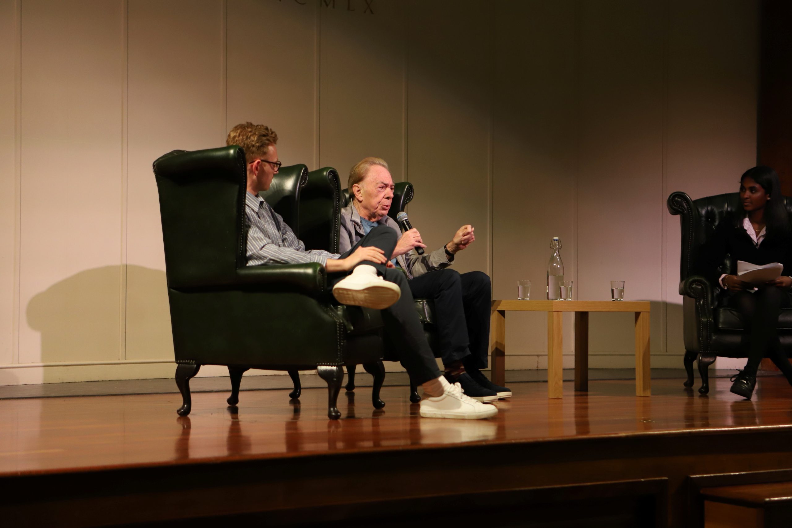 Andrew Lloyd Webber centre stage in an armchair speaking into the mic with his son to his right looking at him - they are on stage with the cream and wood backdrop behind them lit