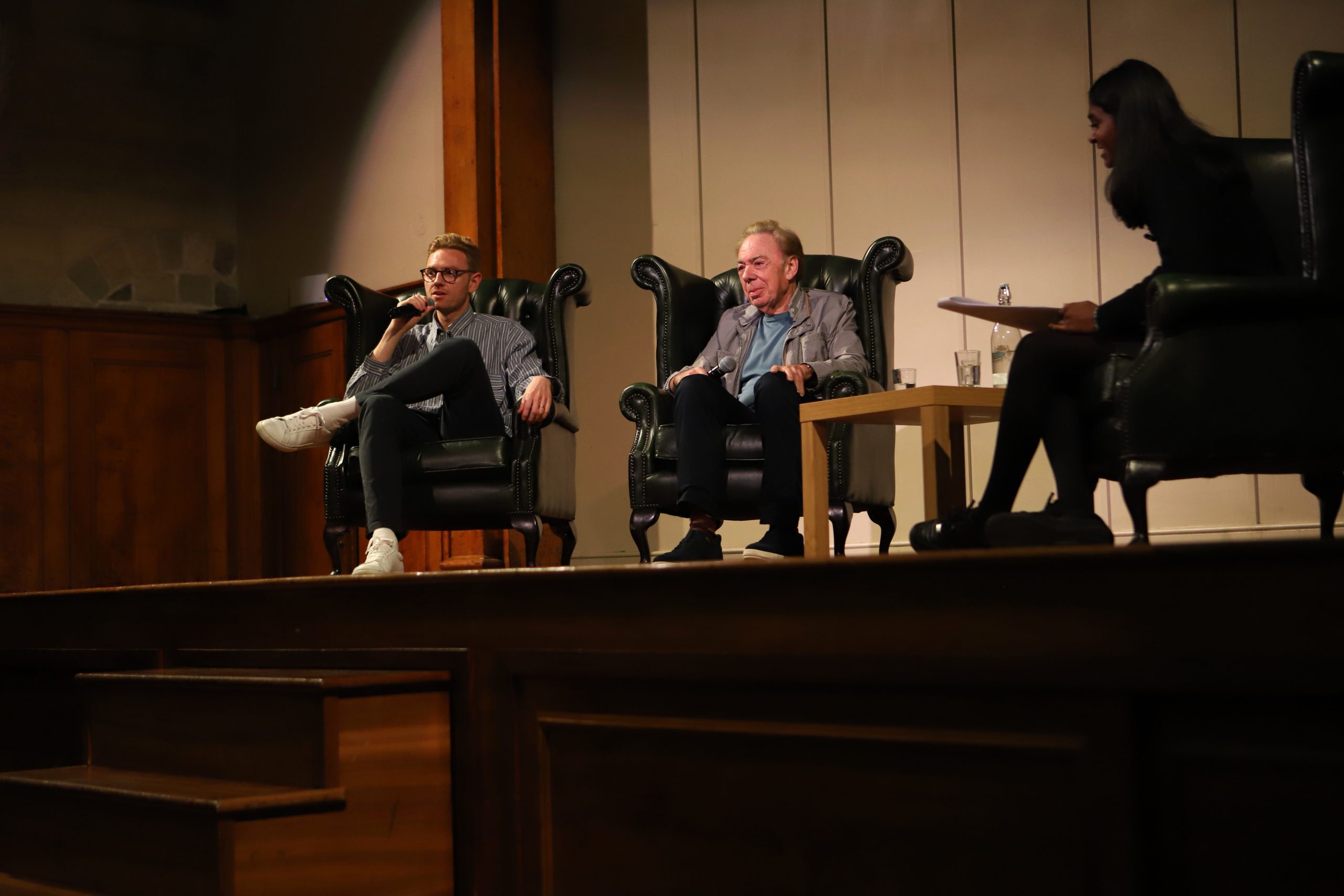 Andrew Lloyd Webber centre stage smiling in an armchair speaking into the mic with his son to his right speaking into a microphone and a silhouette of the back of Saathana in her armchair to his left, far right of the photo - they are on stage with the cream and wood backdrop behind them lit
