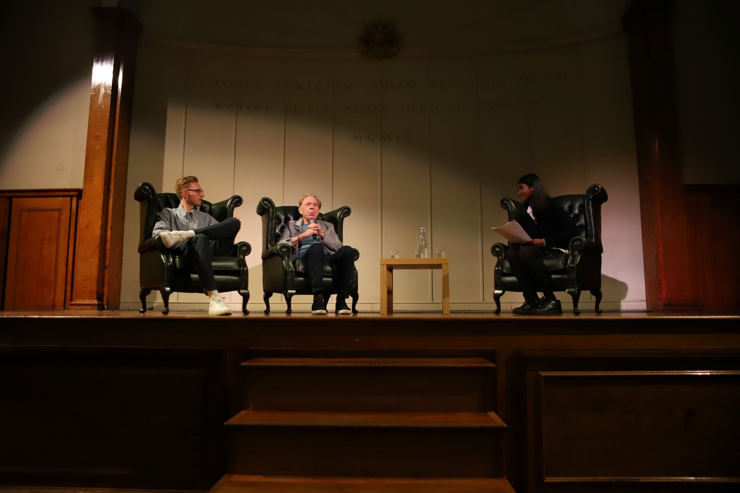 Andrew Lloyd Webber centre stage in an armchair speaking into the mic with his son to his right looking at him and Saathana in her armchair to his left smiling, far right of the photo - they are on stage with the cream and wood backdrop behind them lit