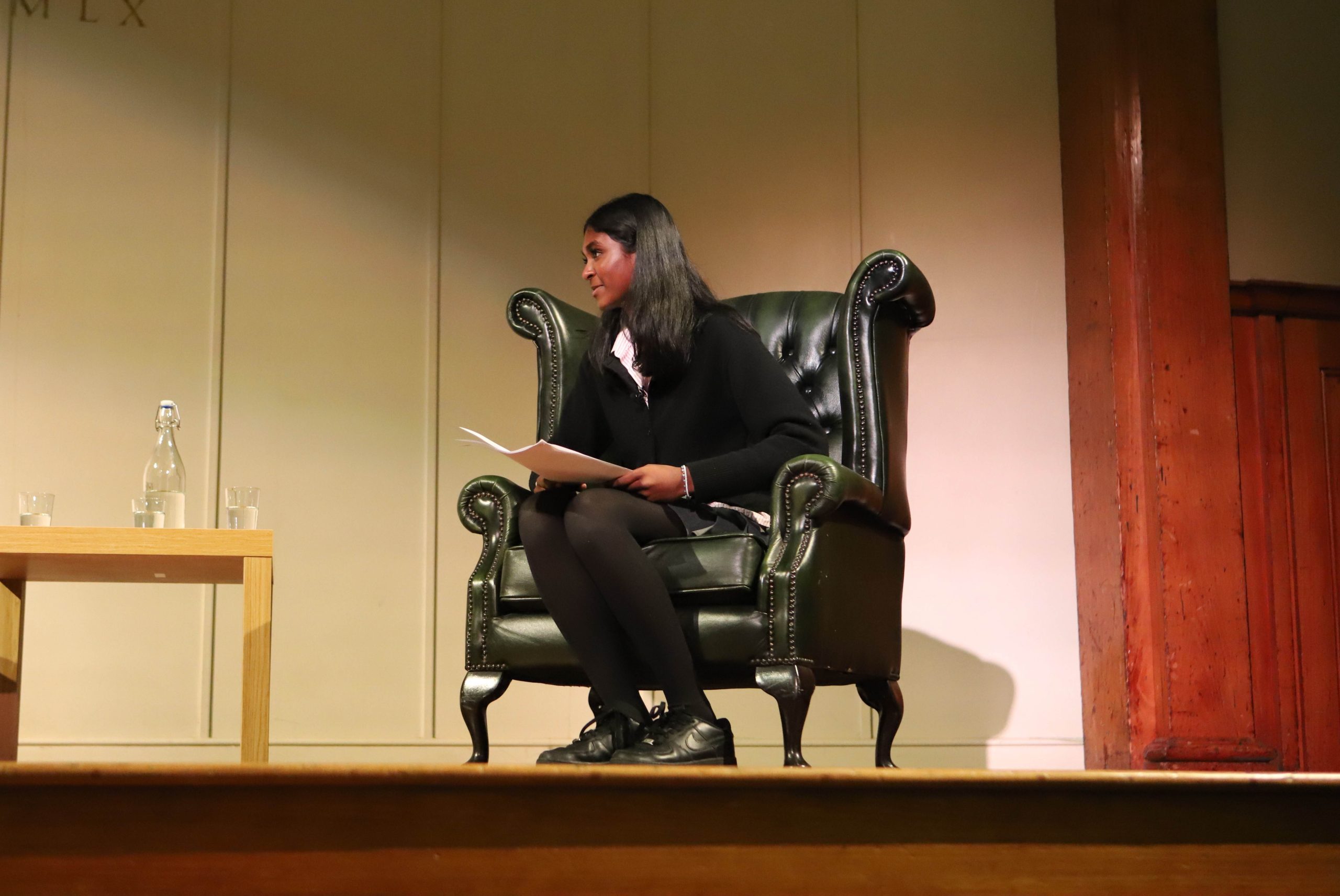 A profile of Saathana sat in a leather armchair on stage with a piece of paper in hand smiling, she is on the wooden stage set against the cream and wooden backdrop