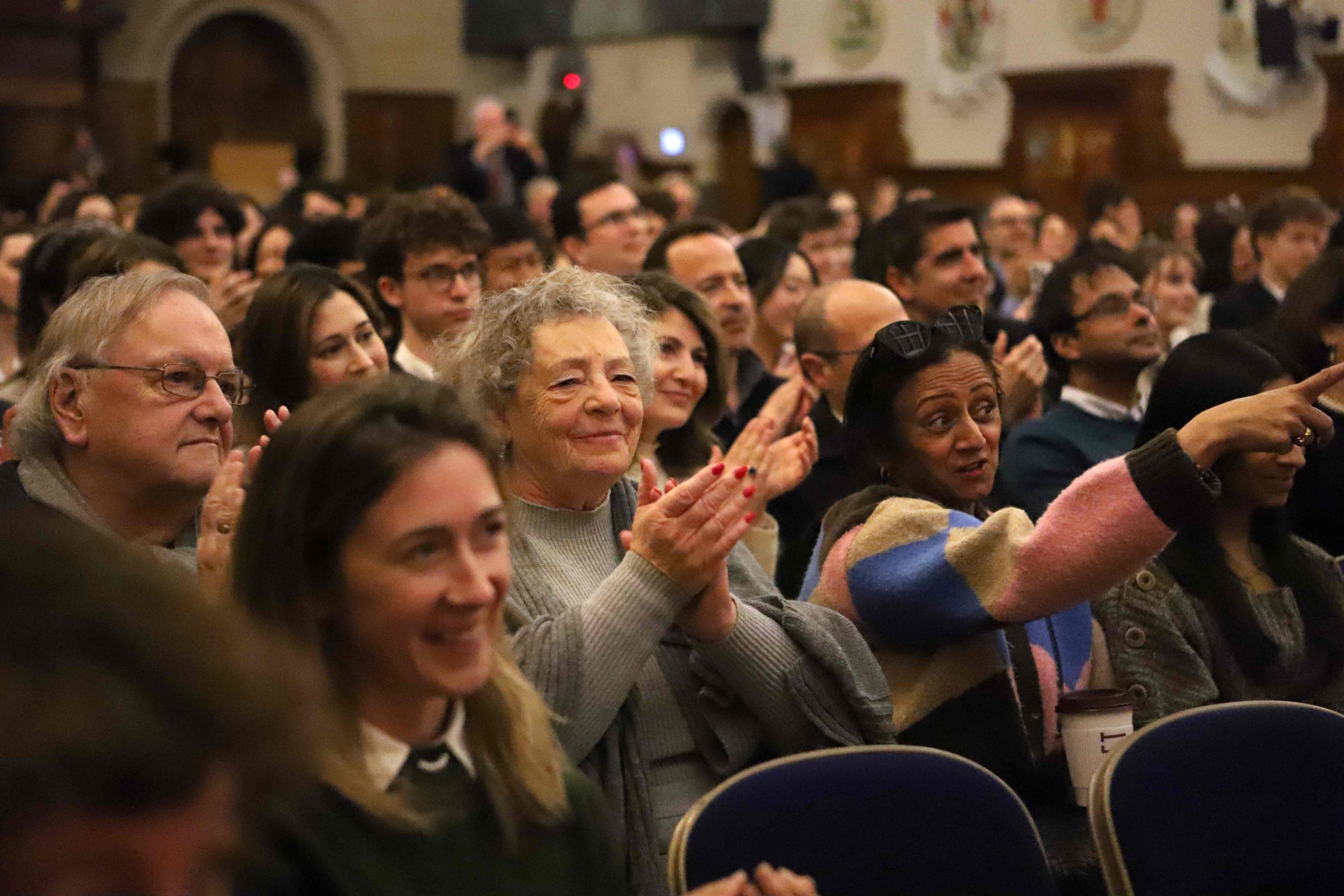 An audience of mostly adults with some pupils dotted around , with wooden panelling and cream walls in the background - everyone is clapping and smiling and one older lady is looking at the camera grinning with the lady next t her pointing forward