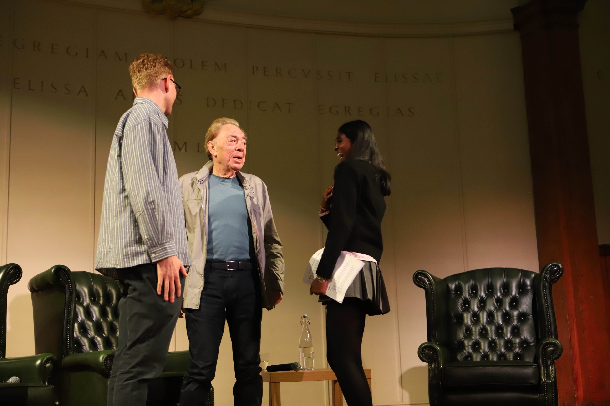 Lloyd Webber and his son Alastair standing on stage talking to Saathana who is laughing at something Lloyd Webber is saying - she is wearing black and he is in a blue t-shirt and grey jacket and dark trousers, his son is wearing a light striped shirt and chinos