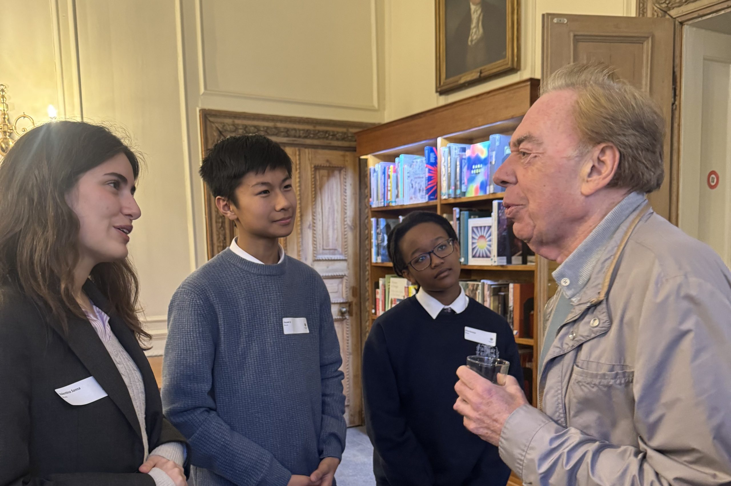 Lloyd Webber to the write speaking to three pupils, two girls and a boy, in the School's library with book shelves in the back ground - the pupils are in a loose line opposite him and the girl on the far left is smiling
