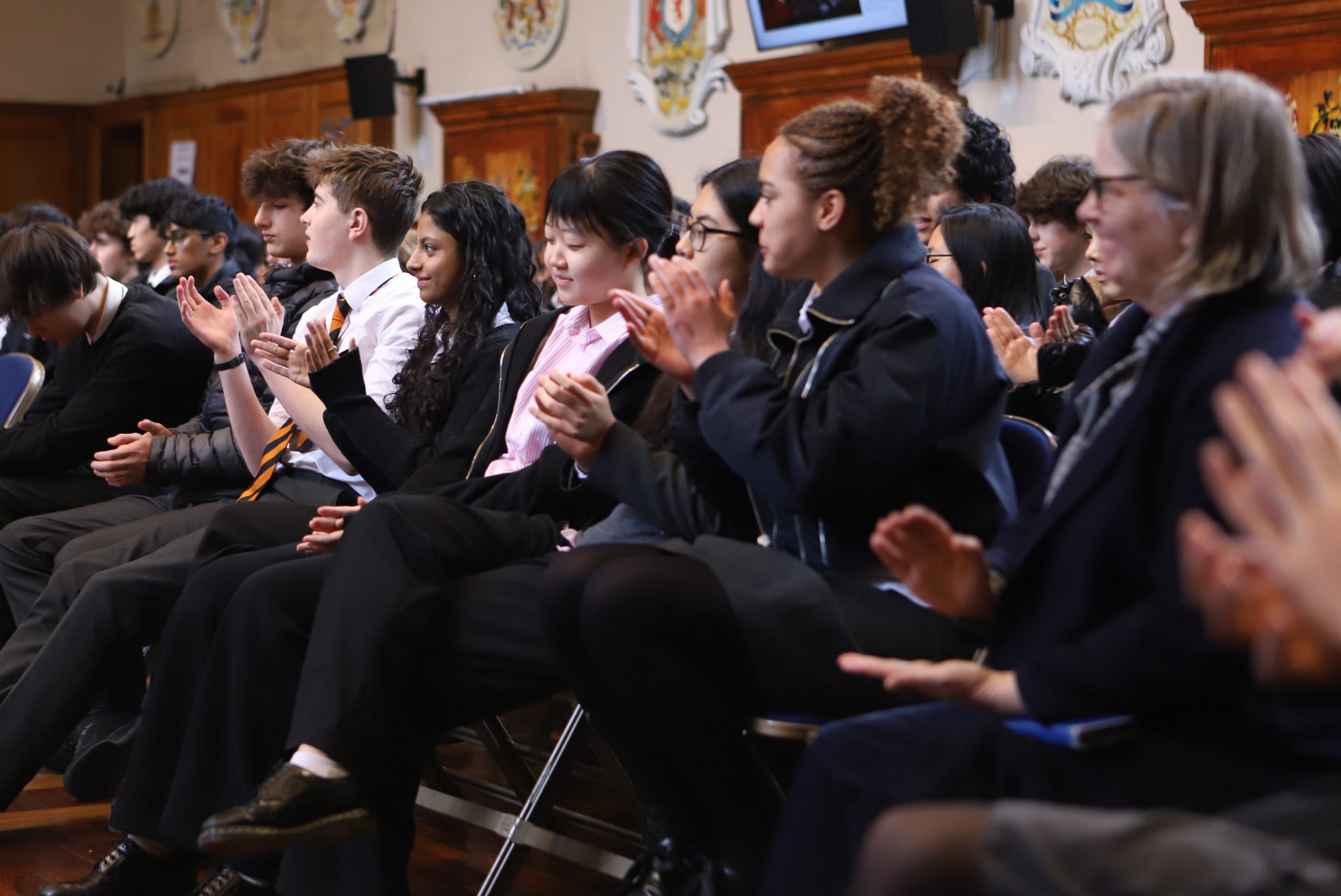 A row of girls and boys clapping and smiling, with wooden panelling and crests on the wall in the background