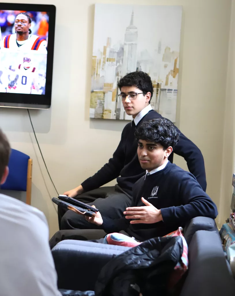 Two boys in Westminster branded sweatshirts, one sat on a couch and the other sat behind him on the arm of the couch, looking at the pupils in front of them, the one in front answering a question with his left hand firmly held up horizontally, with Superbowl images on the screen in the background