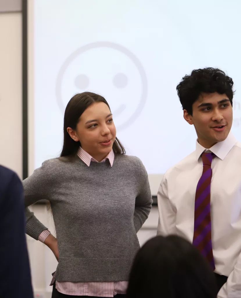 A girl in a grey jumper and a boy in a shirt and tie, standing in front of a screen with a smiley face on, with the backs of two pupils in the foreground showing they are in conversation