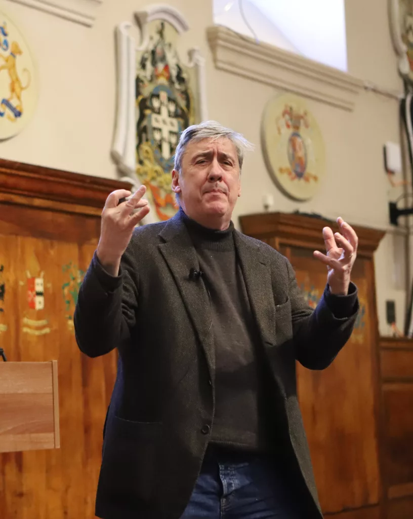 Andrew Graham Dixon standing and speaking, with two hands held up characterfully, with wooden panelling behind him and and crests and logos painted on it