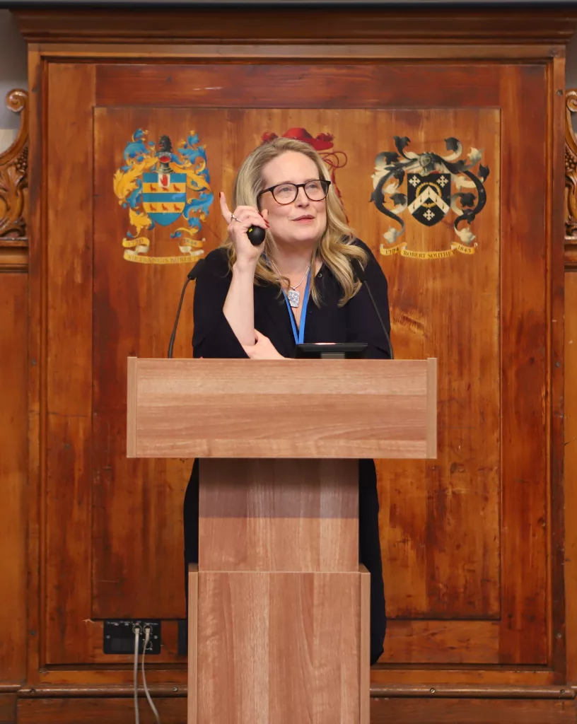 Ophelia Field in the centre, leaning on a podium thoughtfully, using her right elbow, with her finger in the air, with wooden panelling behind her and a crest on it either side of her