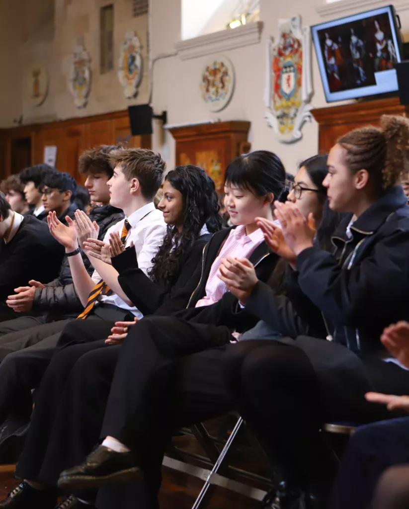 A row of girls and boys clapping and smiling, with wooden panelling on the walls behind them and crests and logos too