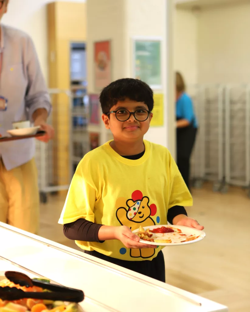A boy in a yellow Children in Need t-shirt with classes smiling at the camera holding his dinner plate of food, with the lunch service area in front and a teacher with a tray behind him