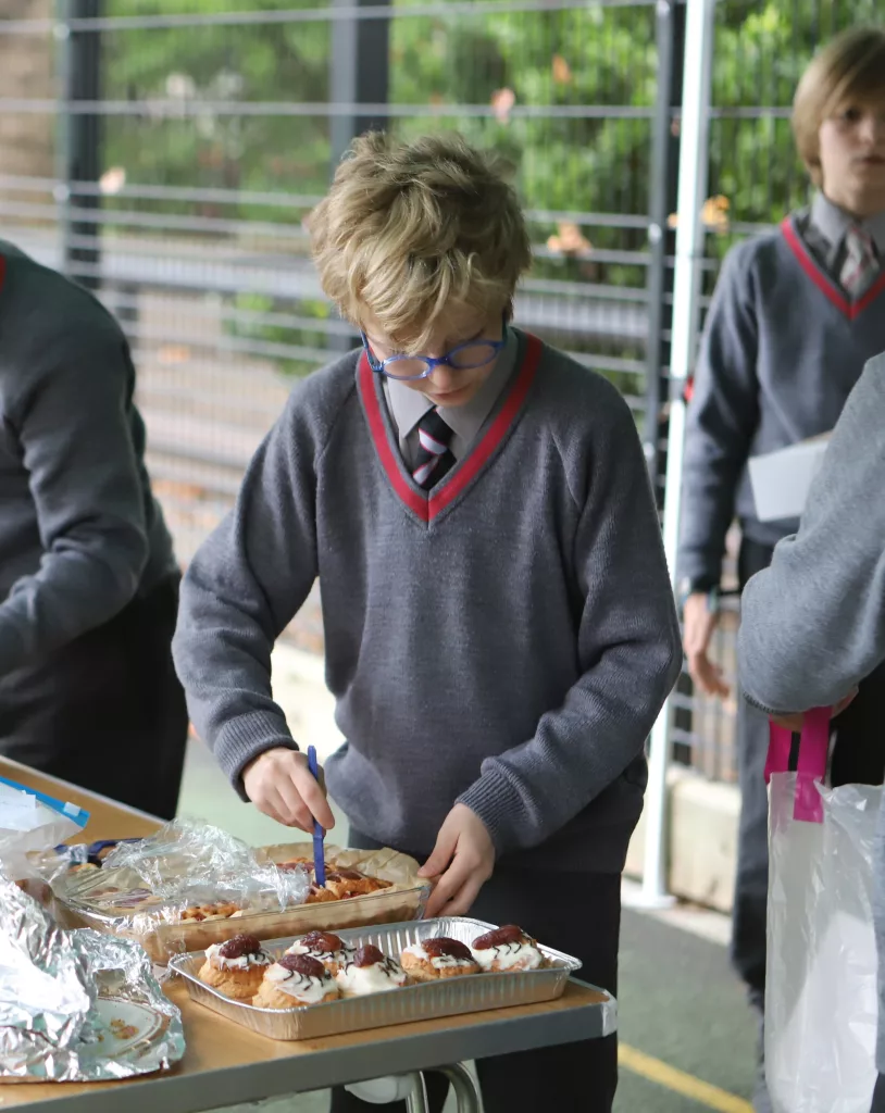 A boy cutting up cupcakes in a box, looking down wearing a grey school jumper with other pupils busy around him and tennis court fences and trees behind him