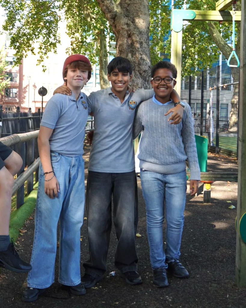 Three boys with their arms around each other in grey school t-shirts with the school logo and jeans, there are playground apparatus to the right and a fence to the left, with trees in the background