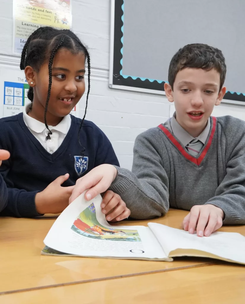 A boy in a grey school jumper sitting at a desk with a girl in a navy uniform on his right, turning the page of a book and looking at it together, with a board in the background