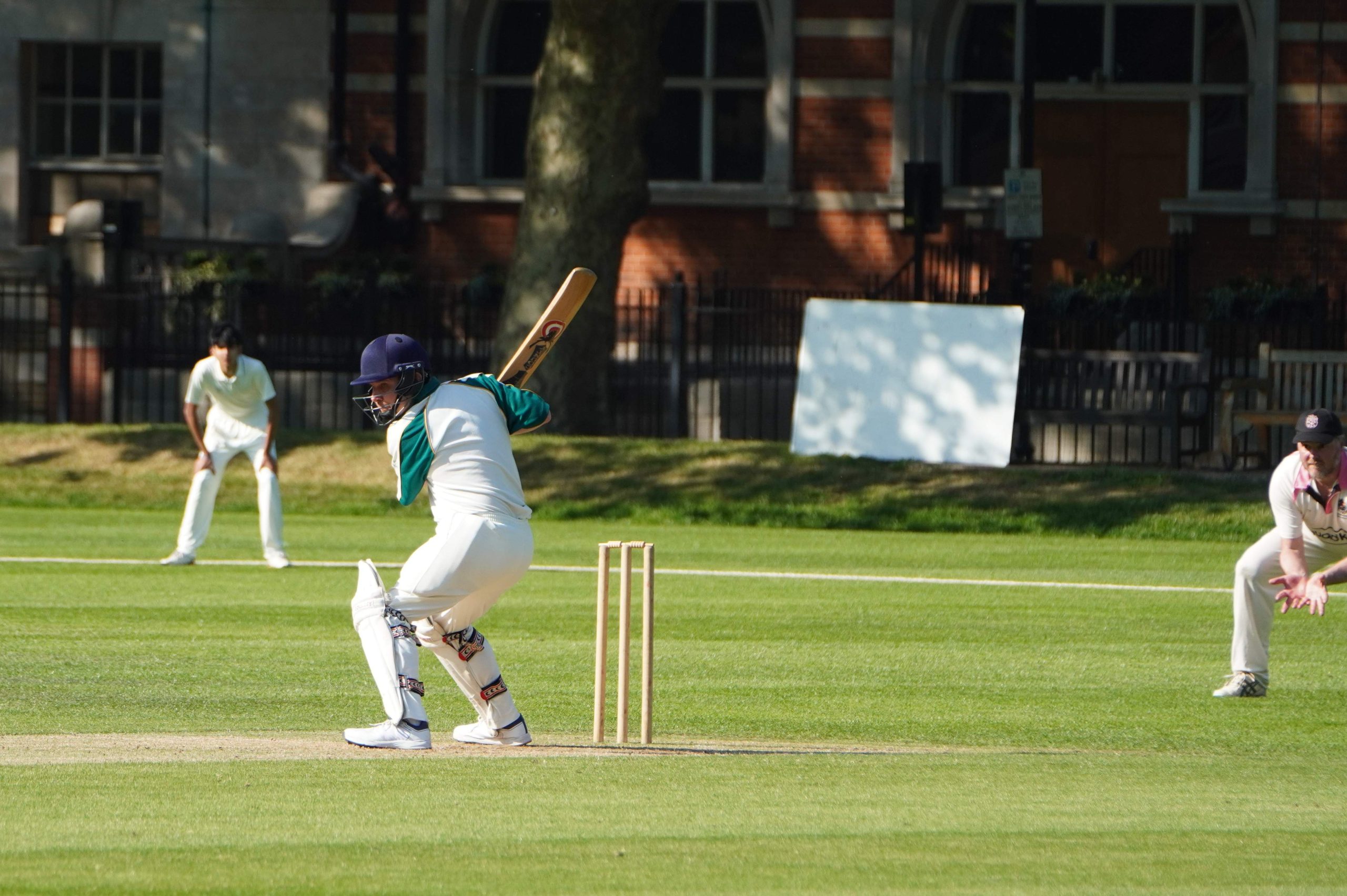 An Author is about to bat, he is wearing a green-and-white top and is in front of the wicket - you can see the score board to the right and a fielder on the left with redbrick building and trees behind them