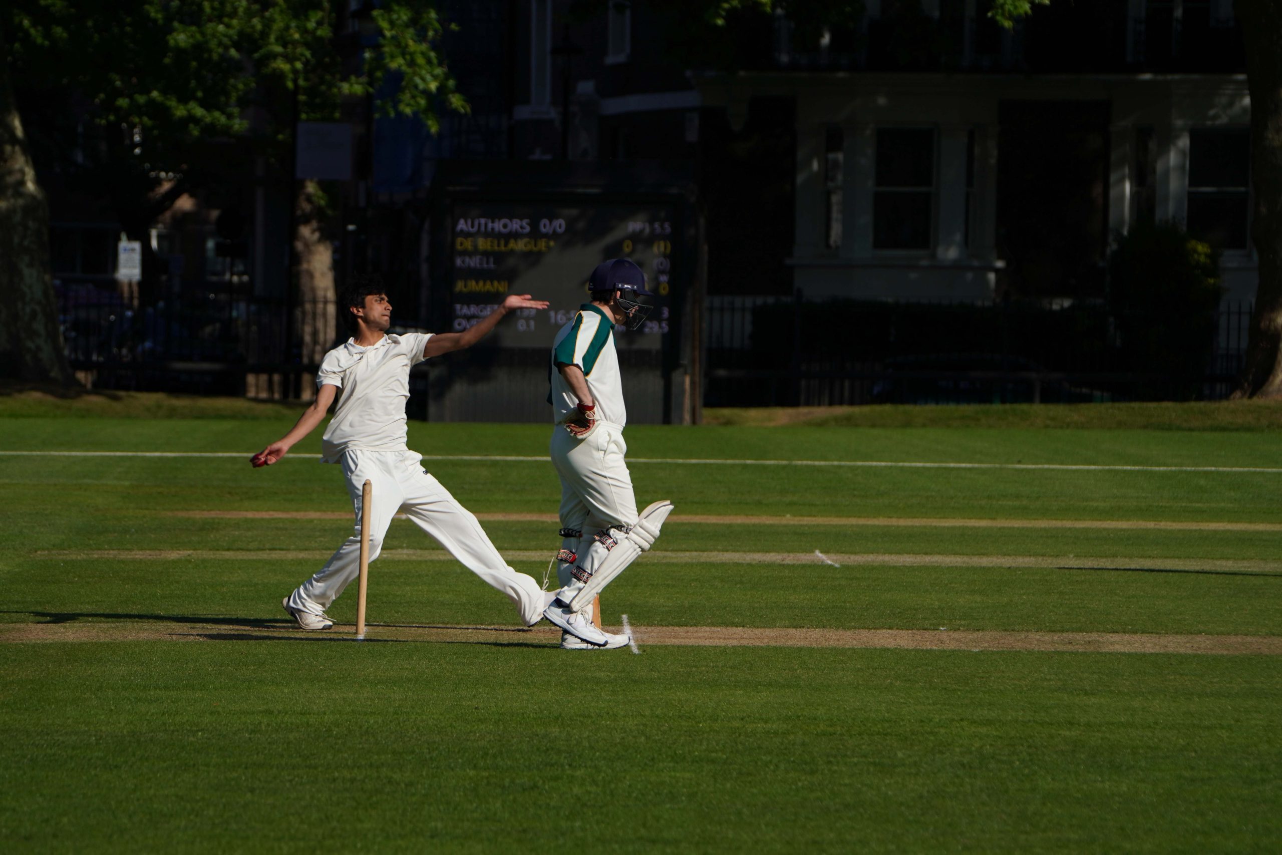 A Westminster pupil bowling, about to swing him arm and there is an author strolling in front of him with leg pads on