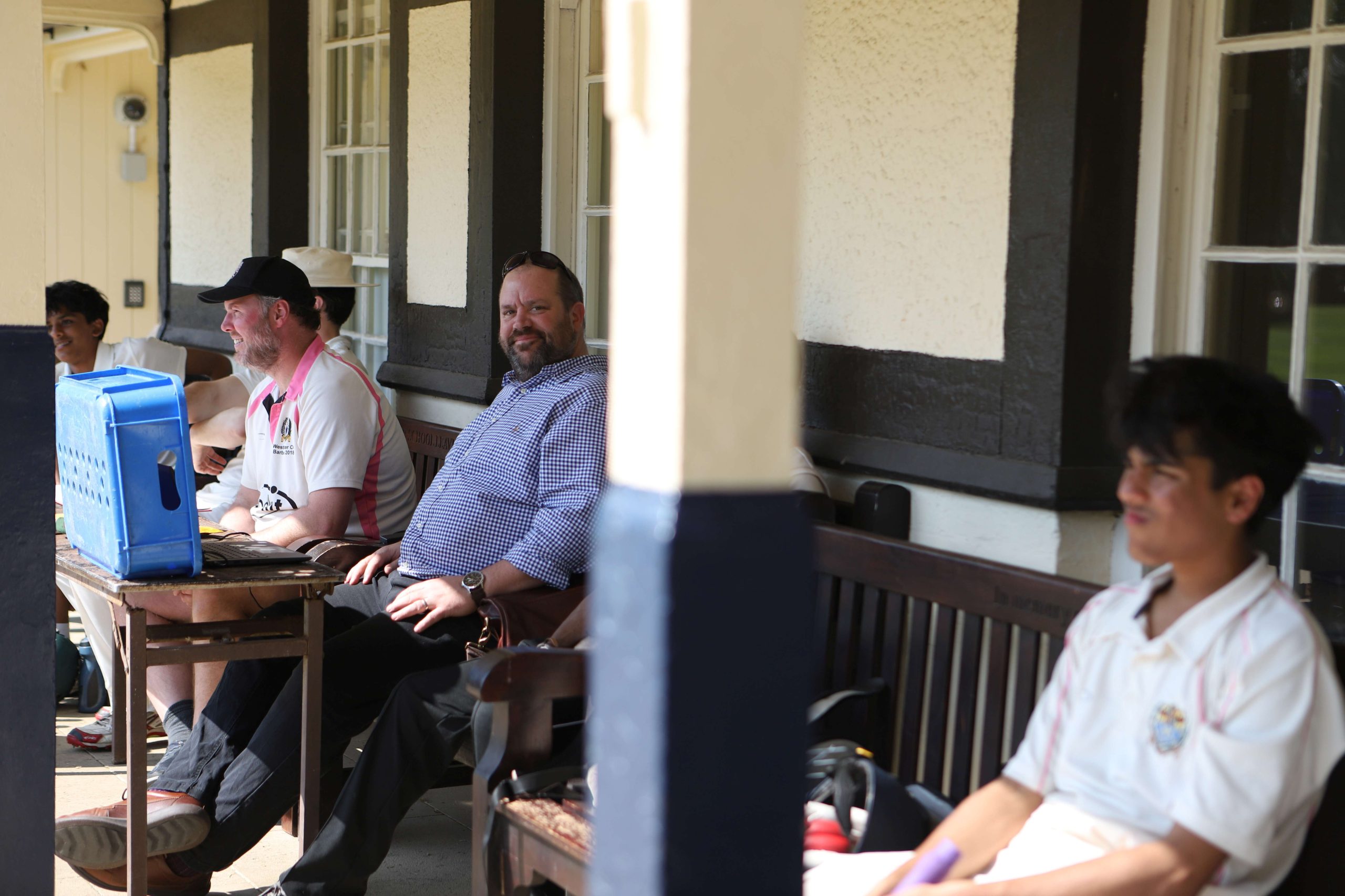 A man in a checked shirt is smiling at the camera, he is sat on a bench next to another member of staff in a pink-and-white Westminster top and cap and they are scoring under the canape of the pavilion