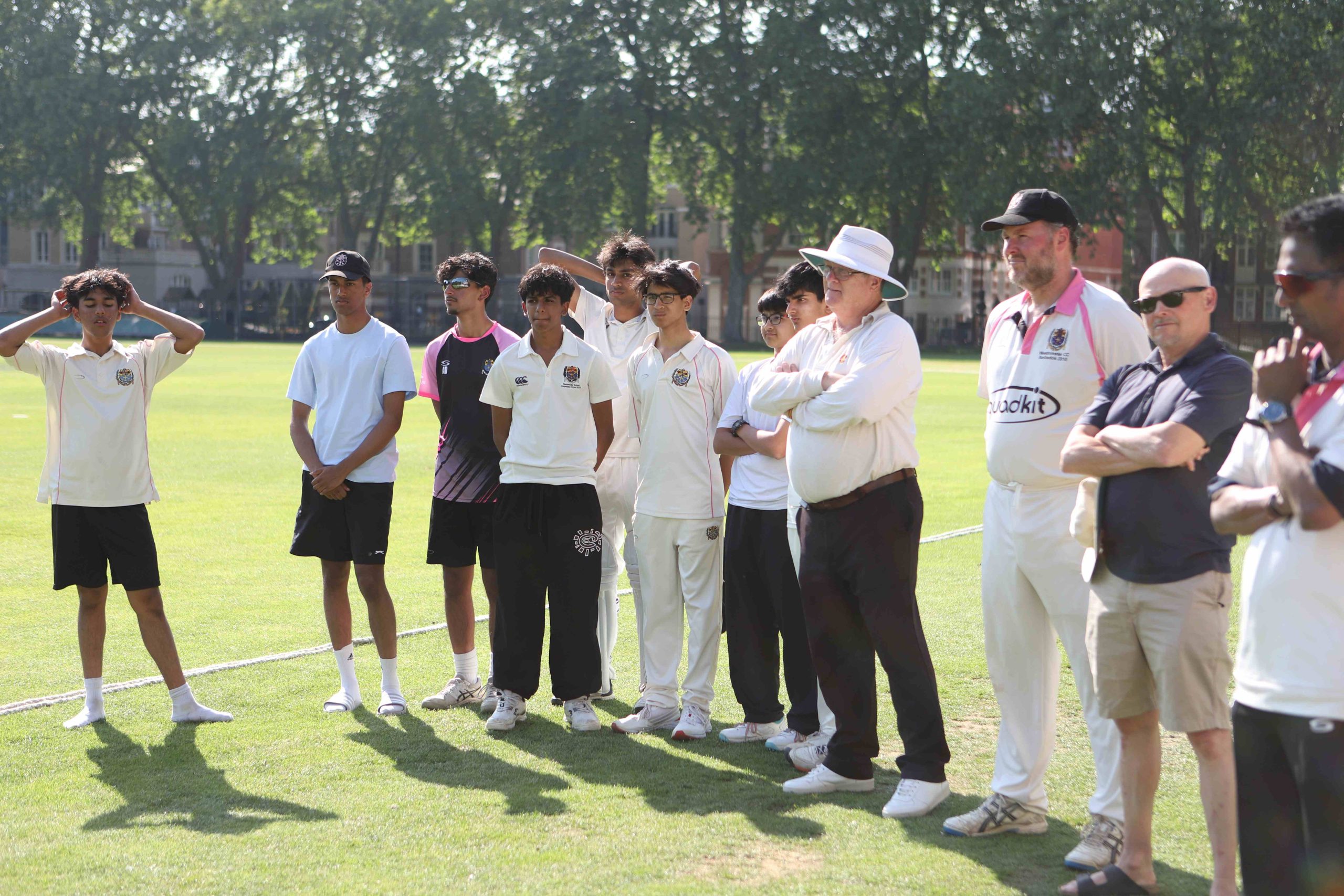 Pupils in cricket whites and Westminster sports clothes in a line on the bright green grass with a line of trees in the background, they are listening to someone speaking