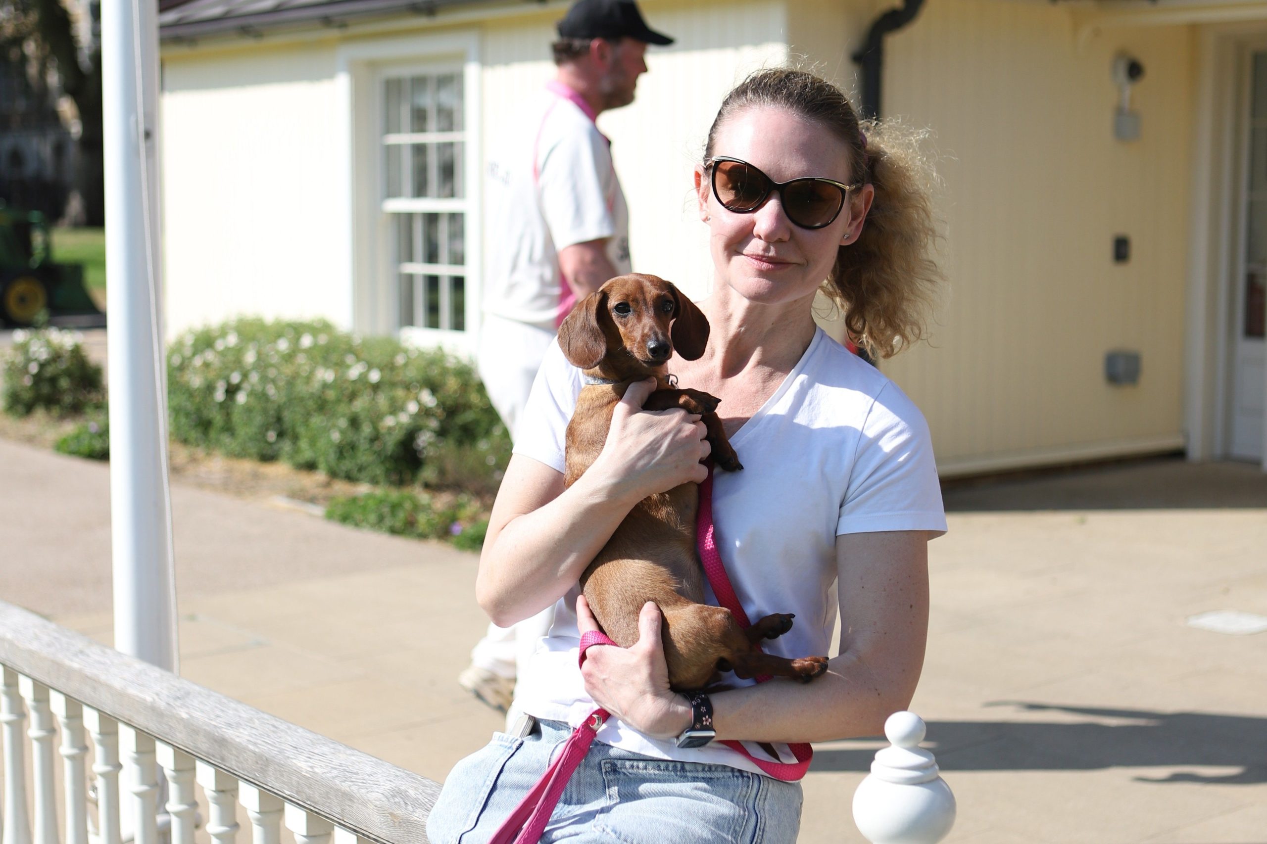 A lady in a white t-shirt holding a dachshund for the camera, she is sat on a white picket fence and the pavilion with white walls is behind her and a cricketer is walking by on the paving