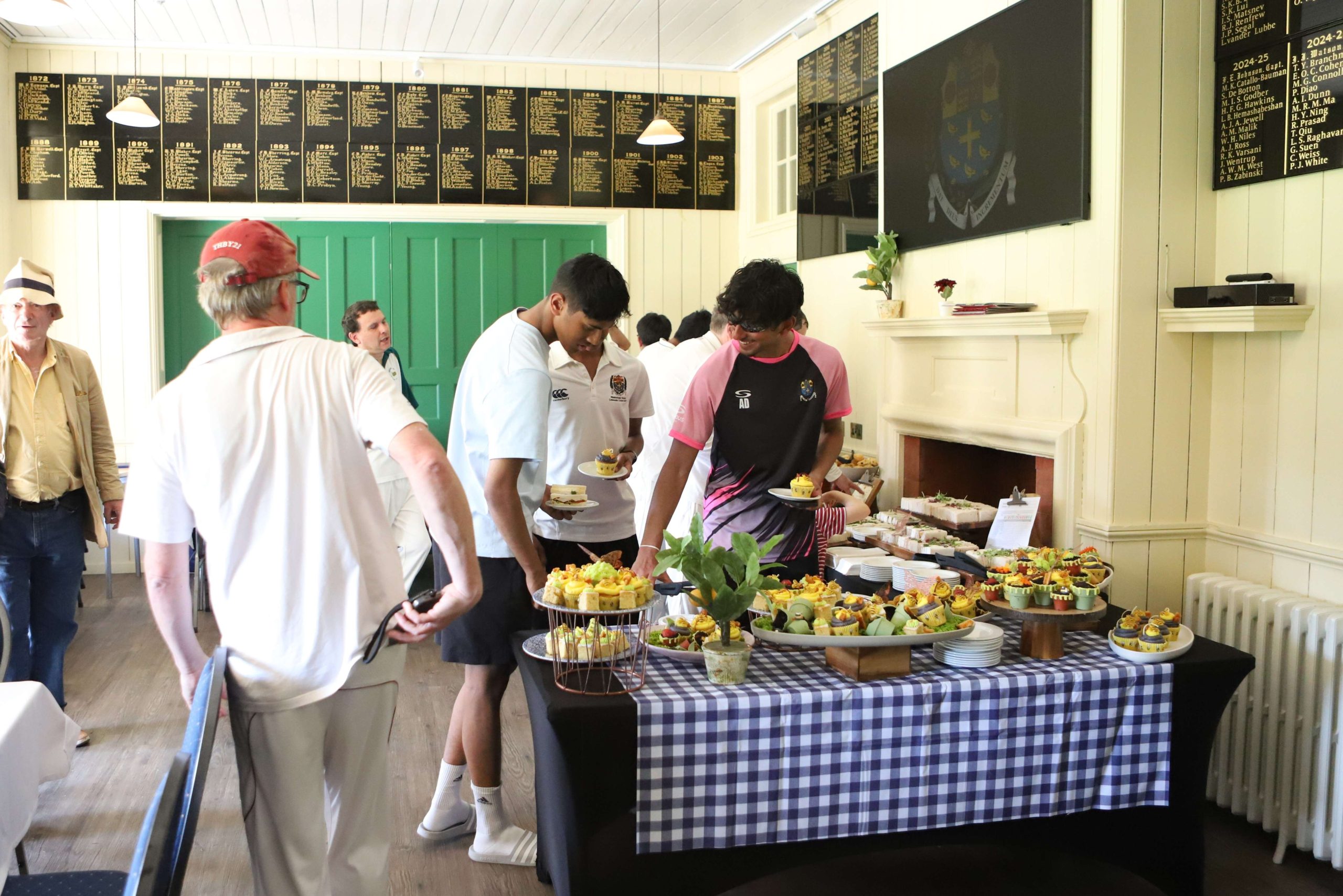 Three Westminster pupil cricketers in sports kit selecting items from the themed buffet of yellow cakes and sandwiches, they are smiling and talking - the table has a blue-and-white chequer cloth and there are other people chatting against the aqua green internal wooden doors behind them