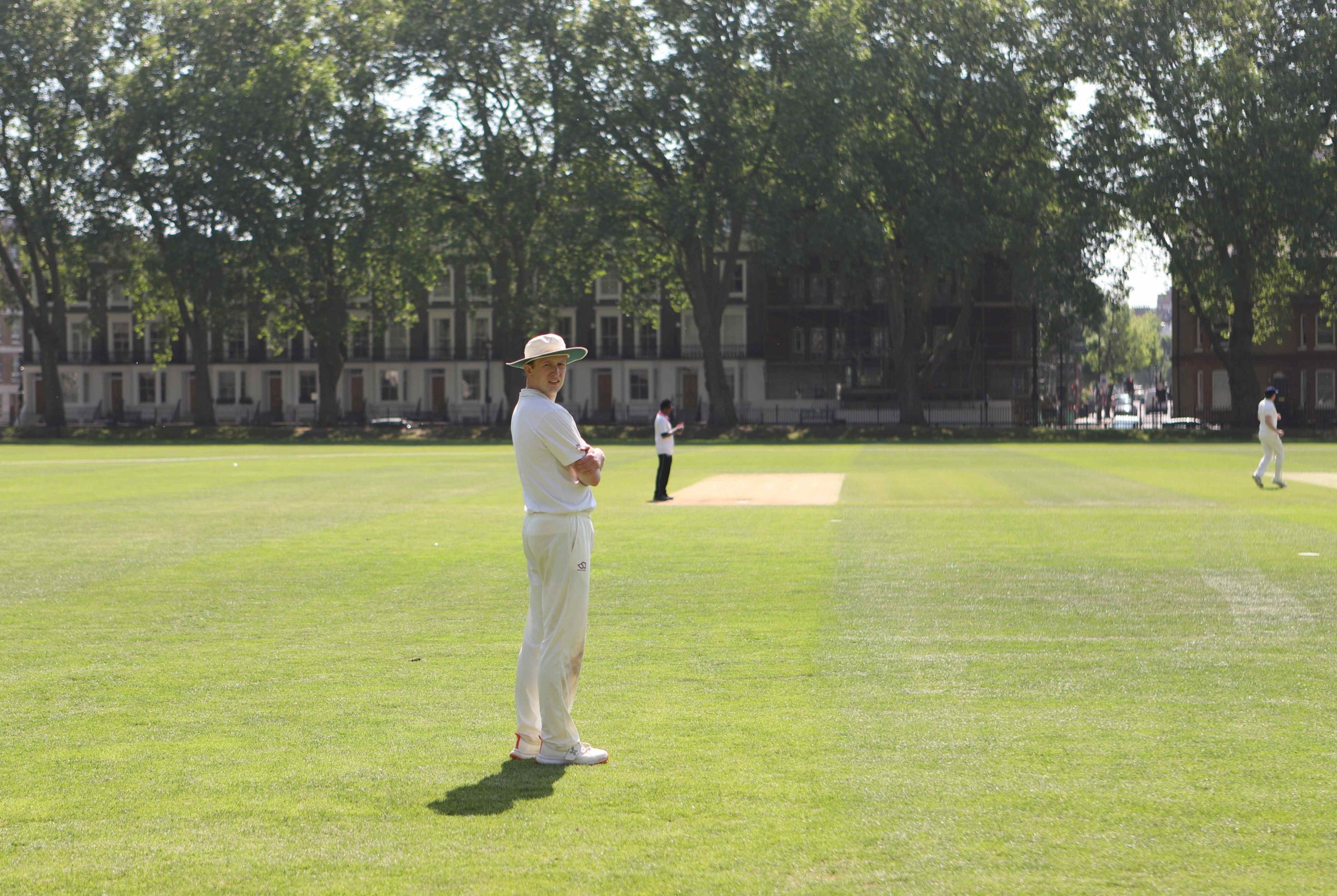 A man standing on the cricket pitch looking back at the camera with his arms folded, he is in whites and there is an umpire in the distance behind him, and behind him are tall redbrick townhouses and trees